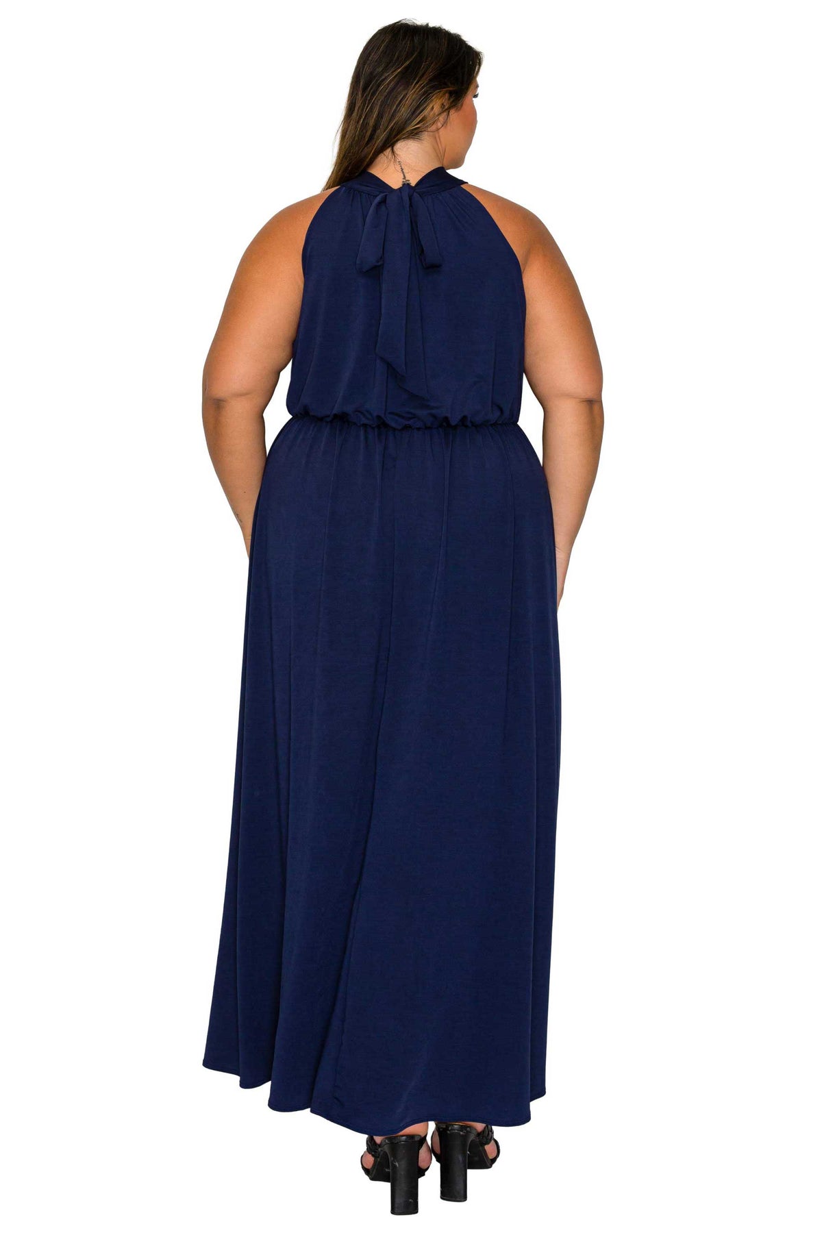 A woman viewed from the back wearing a sleeveless navy maxi dress with a ruffled tie neckline and gathered waist, paired with black heels, against a plain white background.