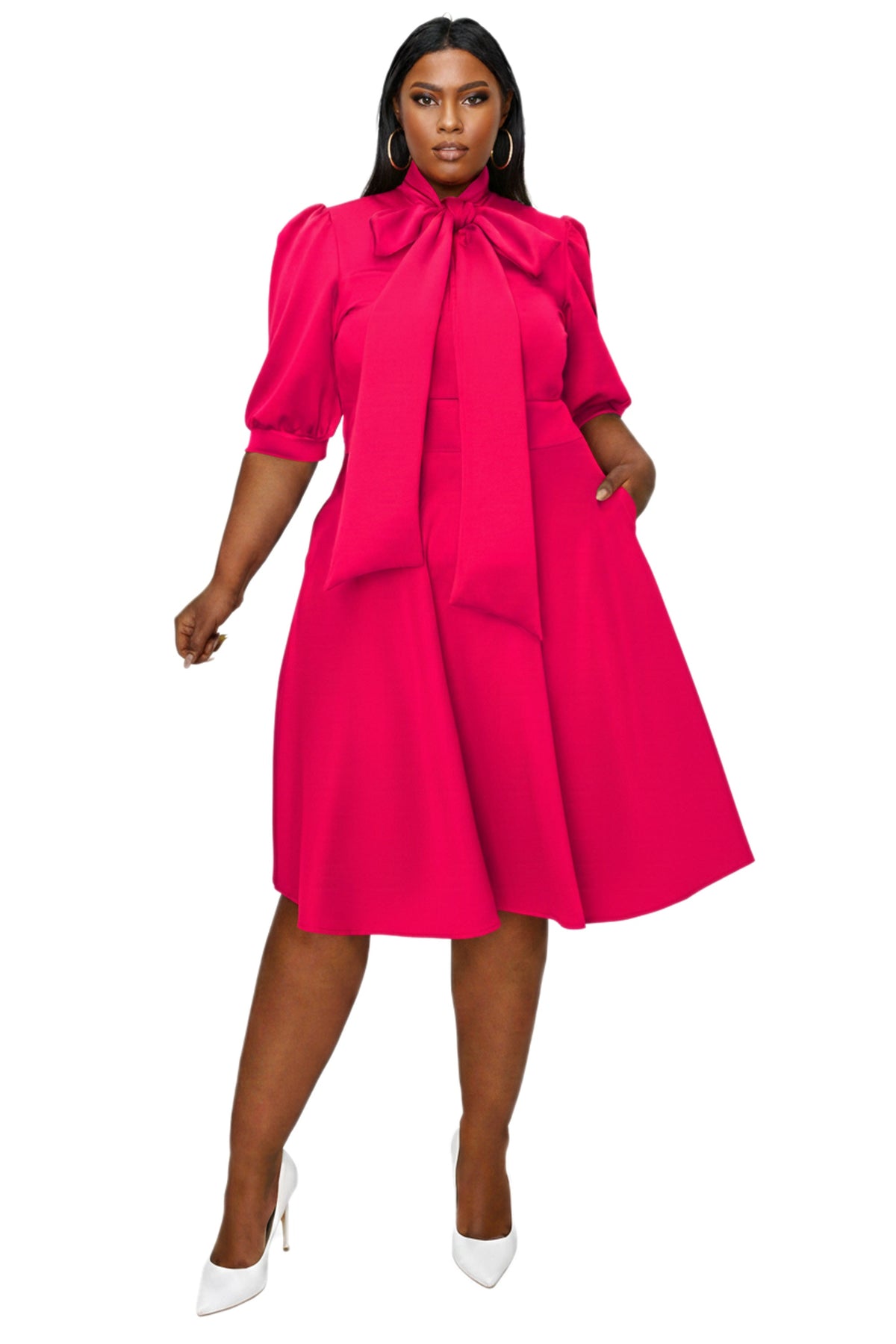 A woman modeling a bright fuchsia pink midi dress with puff sleeves, a large bow at the collar, and a flared skirt, paired with white pointed-toe heels against a plain white background.