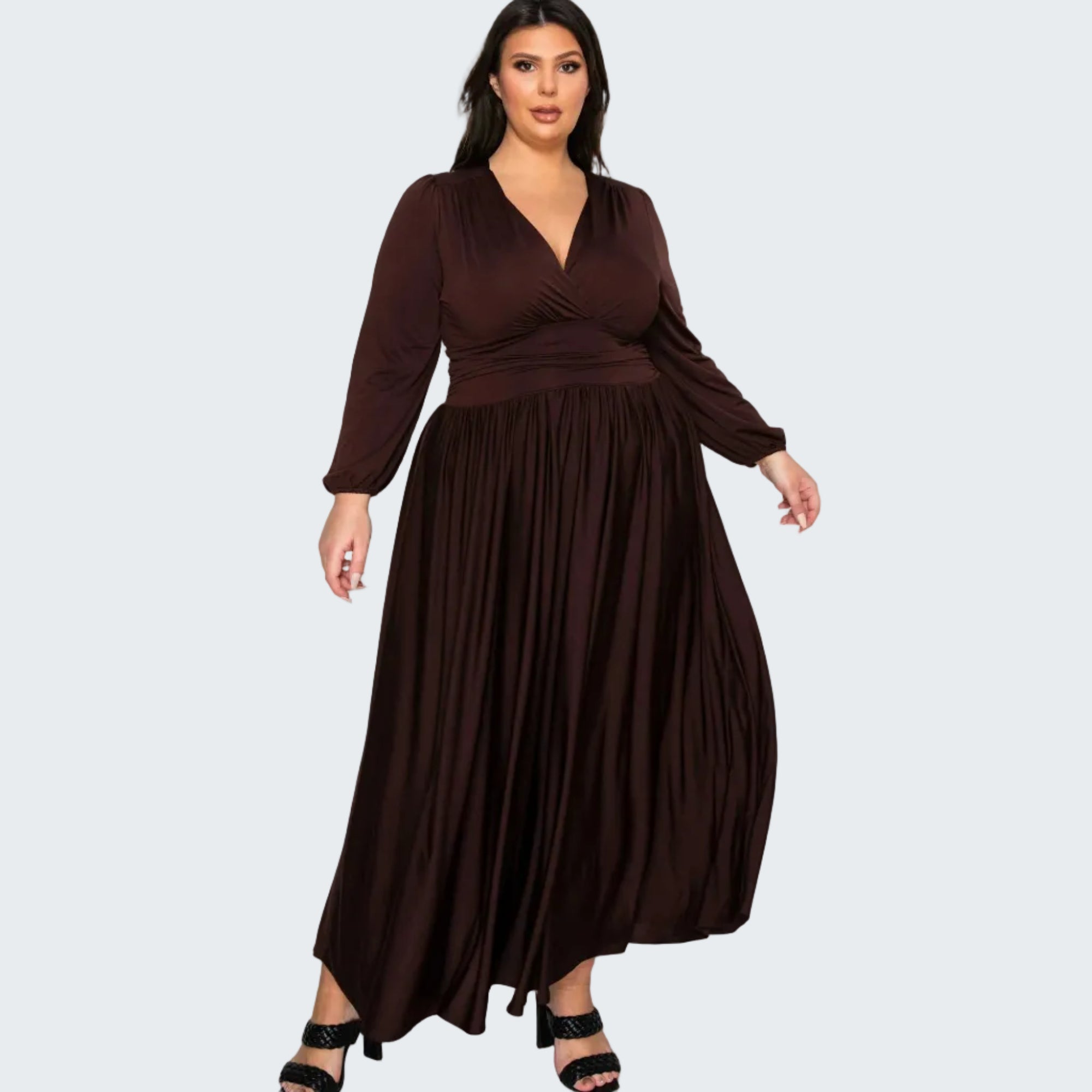 A woman modeling a long-sleeve, wrap-style maxi dress in deep chocolate brown with a pleated skirt, paired with black strappy heels, against a plain white background.