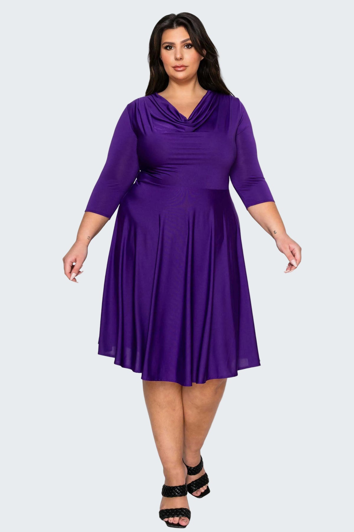 A woman modeling a purple plus-size cowl neck midi dress with three-quarter sleeves and a pleated skirt, paired with black strappy heels, against a solid white background.