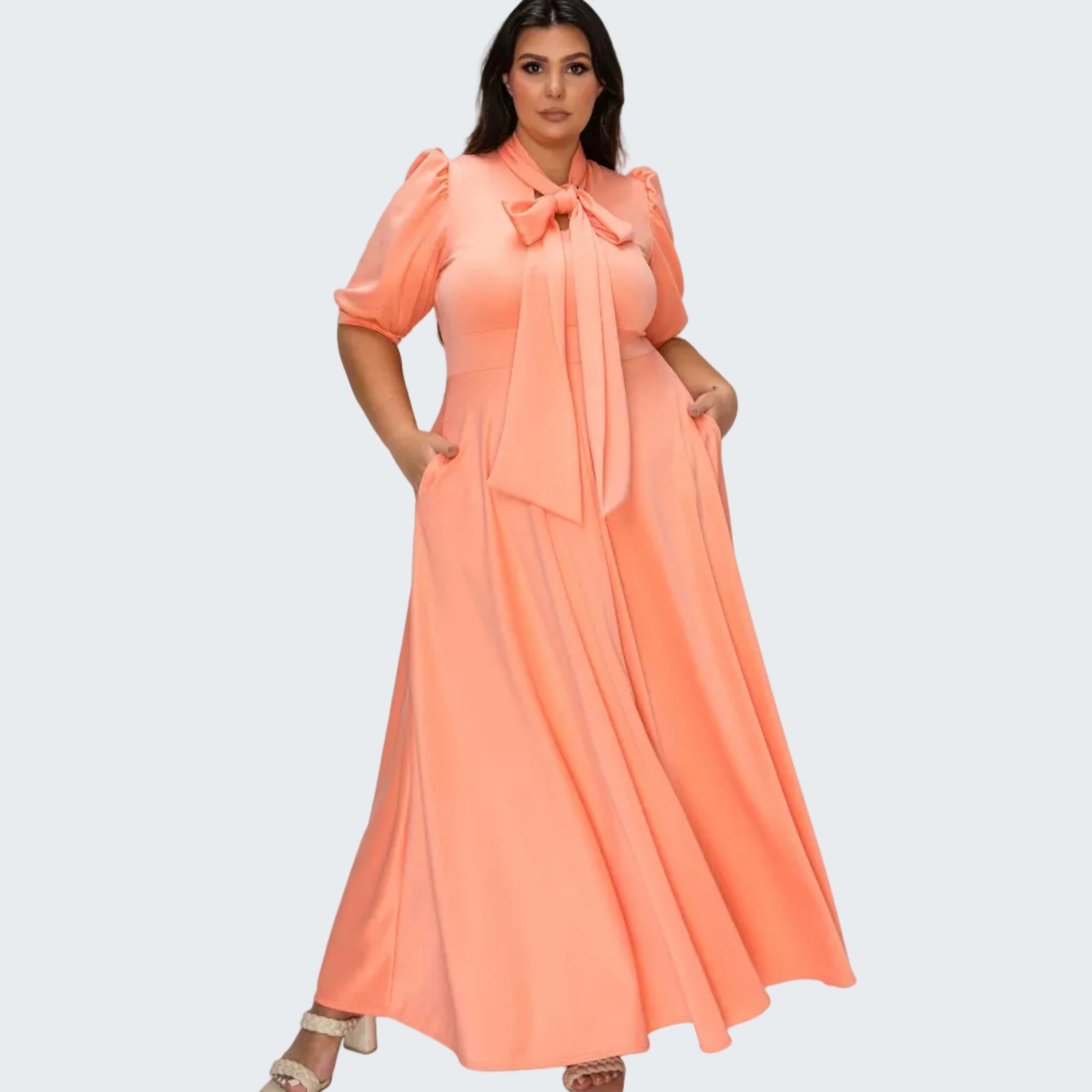 A woman modeling a peach-colored maxi dress with puff sleeves, a large bow at the collar, and side pockets, set against a plain white background.