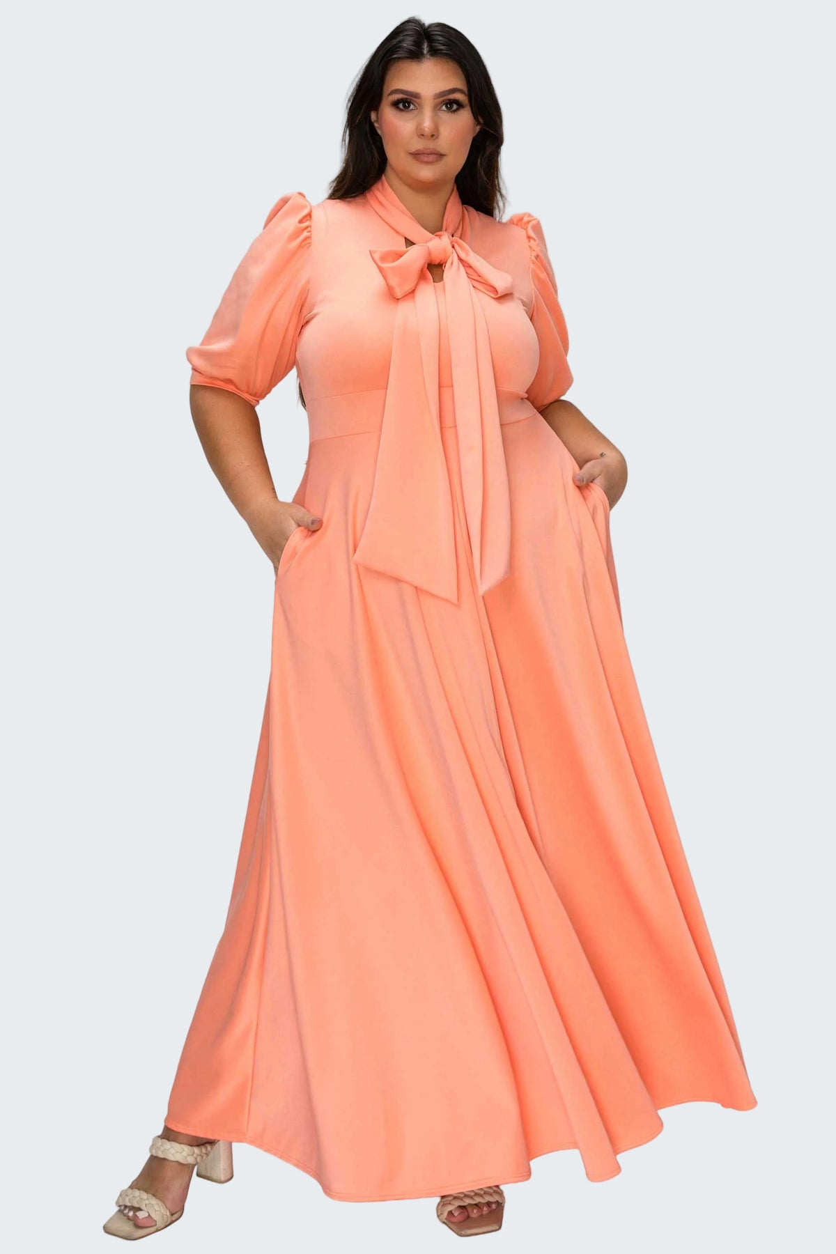 A woman modeling a peach-colored maxi dress with puff sleeves, a large bow at the collar, and side pockets, set against a plain white background.