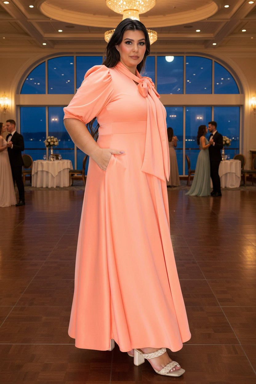 A woman modeling a peach-colored maxi dress with short puff sleeves, a high neck with a bow detail, and side pockets, set against a plain white background.