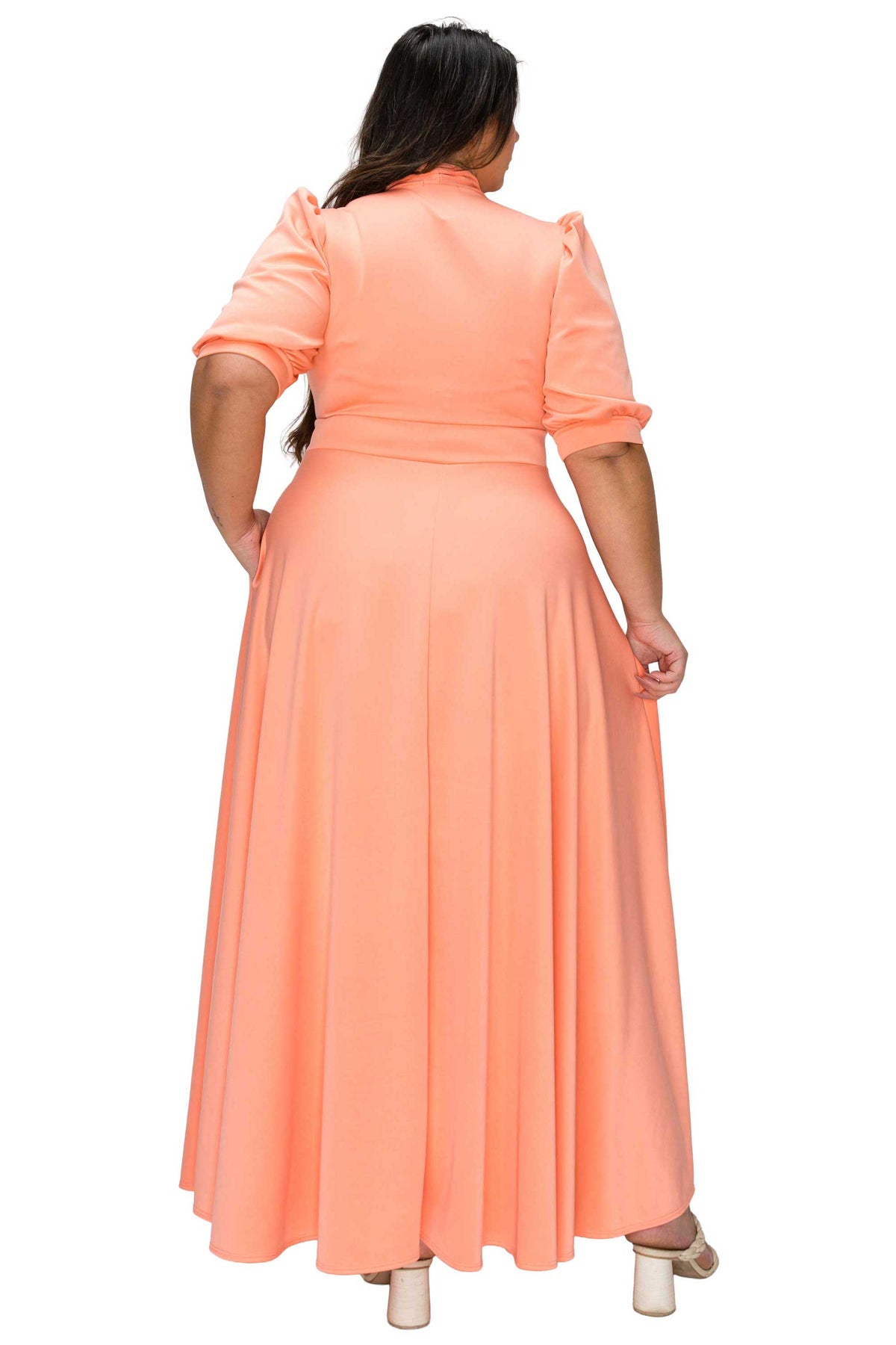 A woman viewed from the back wearing a peach-colored maxi dress with short puff sleeves and a high neckline, paired with beige block-heel sandals, against a plain white background.