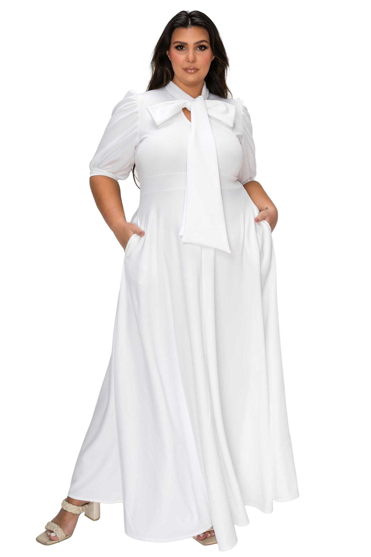 A woman modeling a white maxi dress with short puff sleeves, a large bow collar, and side pockets, paired with beige heeled sandals against a plain white background.