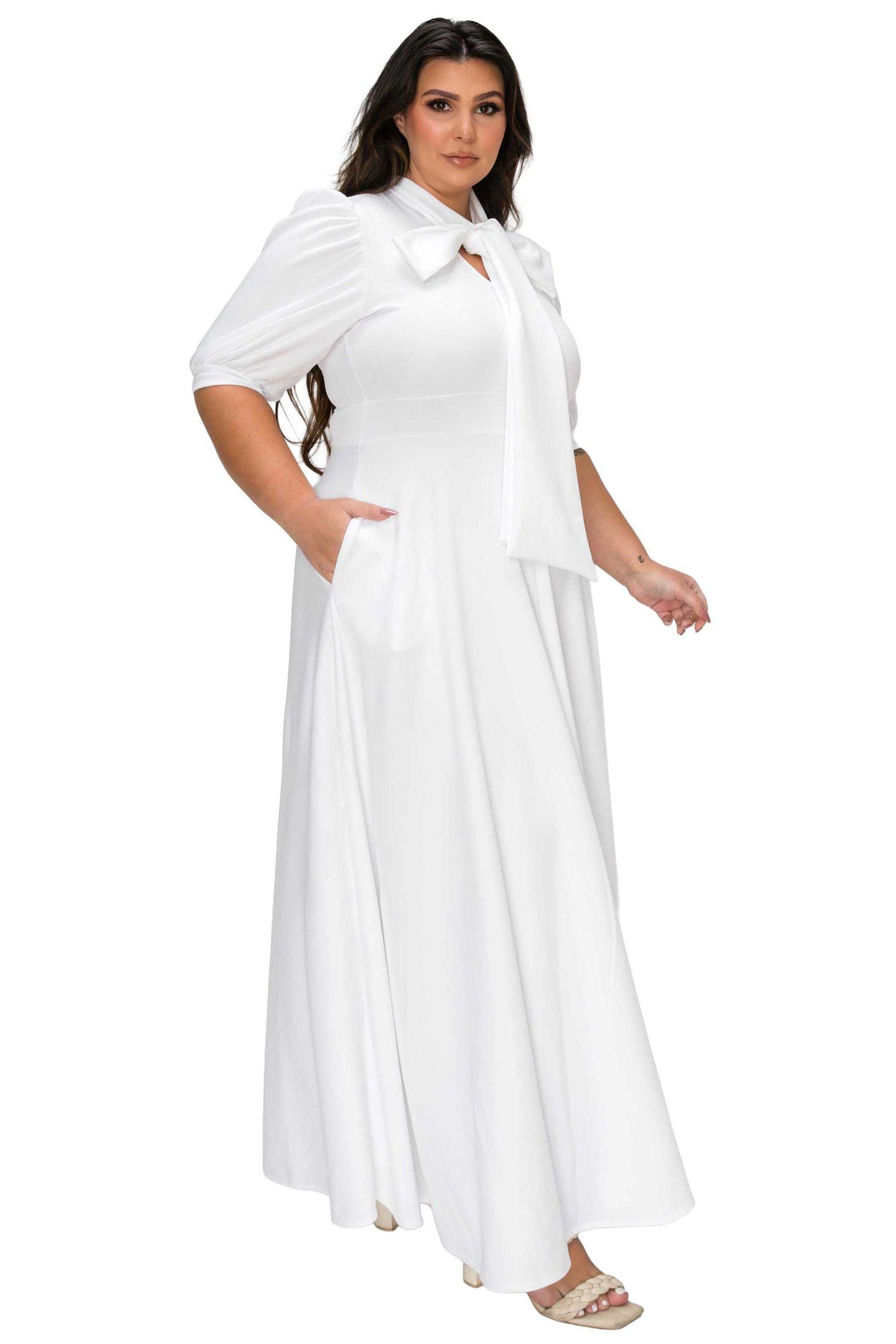 A woman modeling a white maxi dress with short puff sleeves, a large bow at the collar, and side pockets, shown against a plain white background.