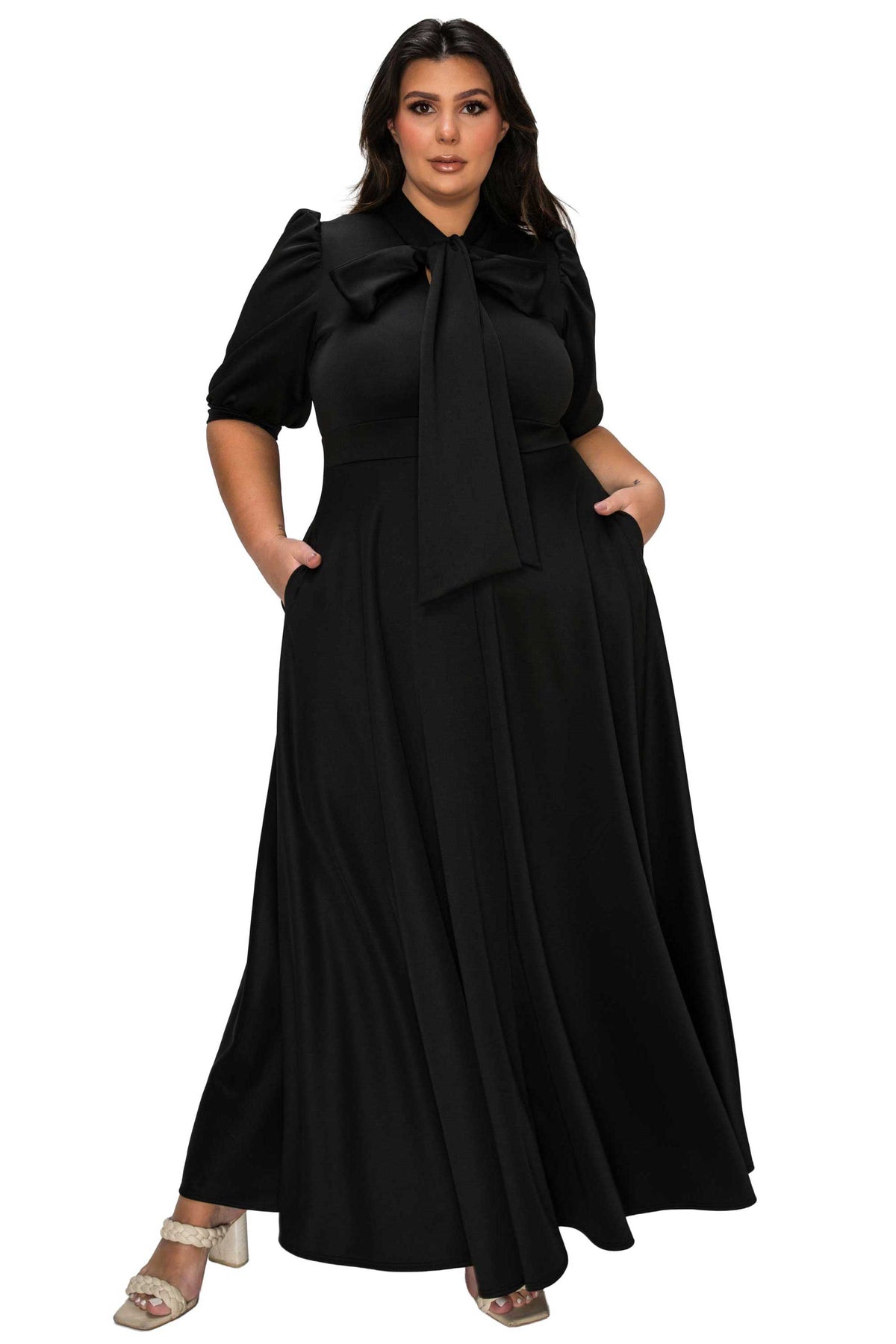A woman modeling a long black maxi dress with short puff sleeves, a large bow at the collar, side pockets, and a flowing skirt, shown against a plain white background.