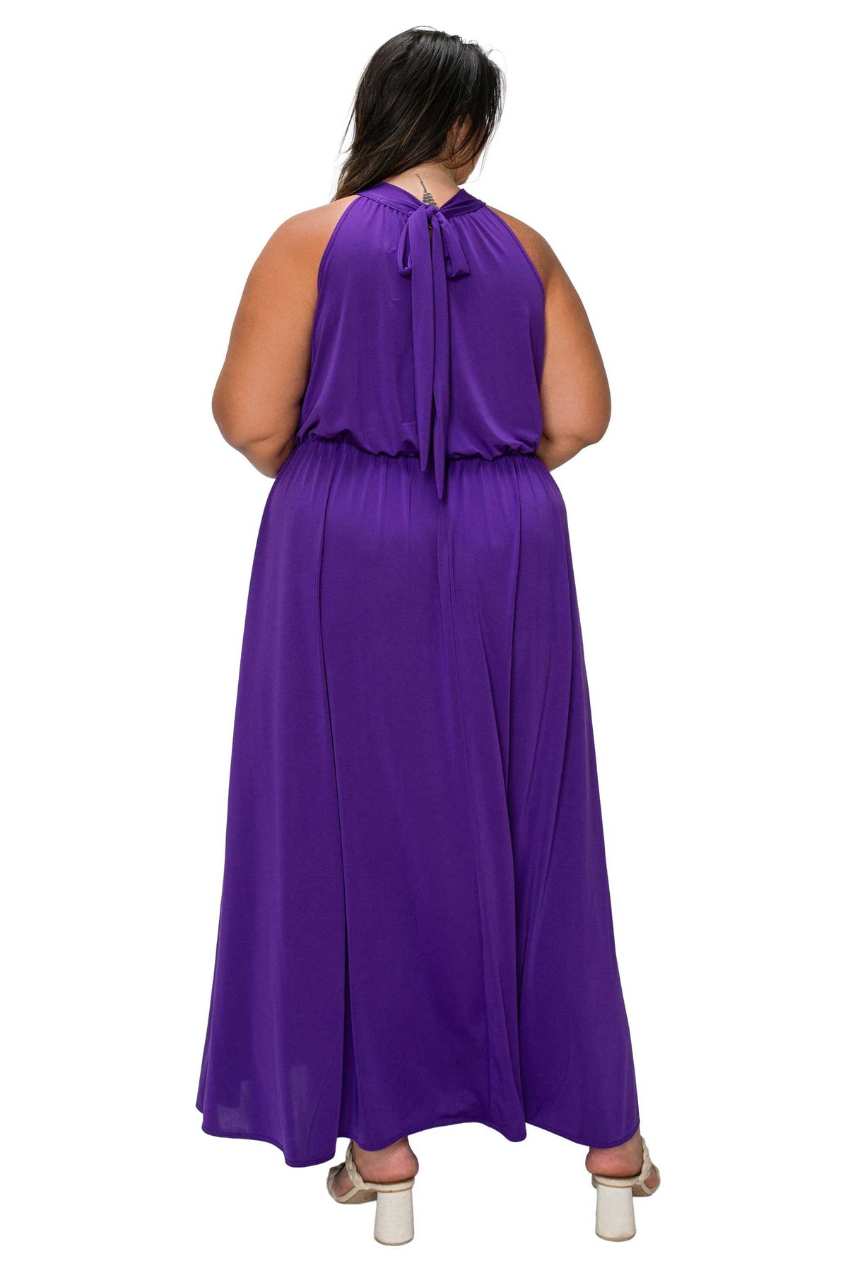 A woman viewed from the back wearing a sleeveless purple maxi dress with a tie-neck detail and a flowing skirt, paired with white heeled sandals, against a plain white background.