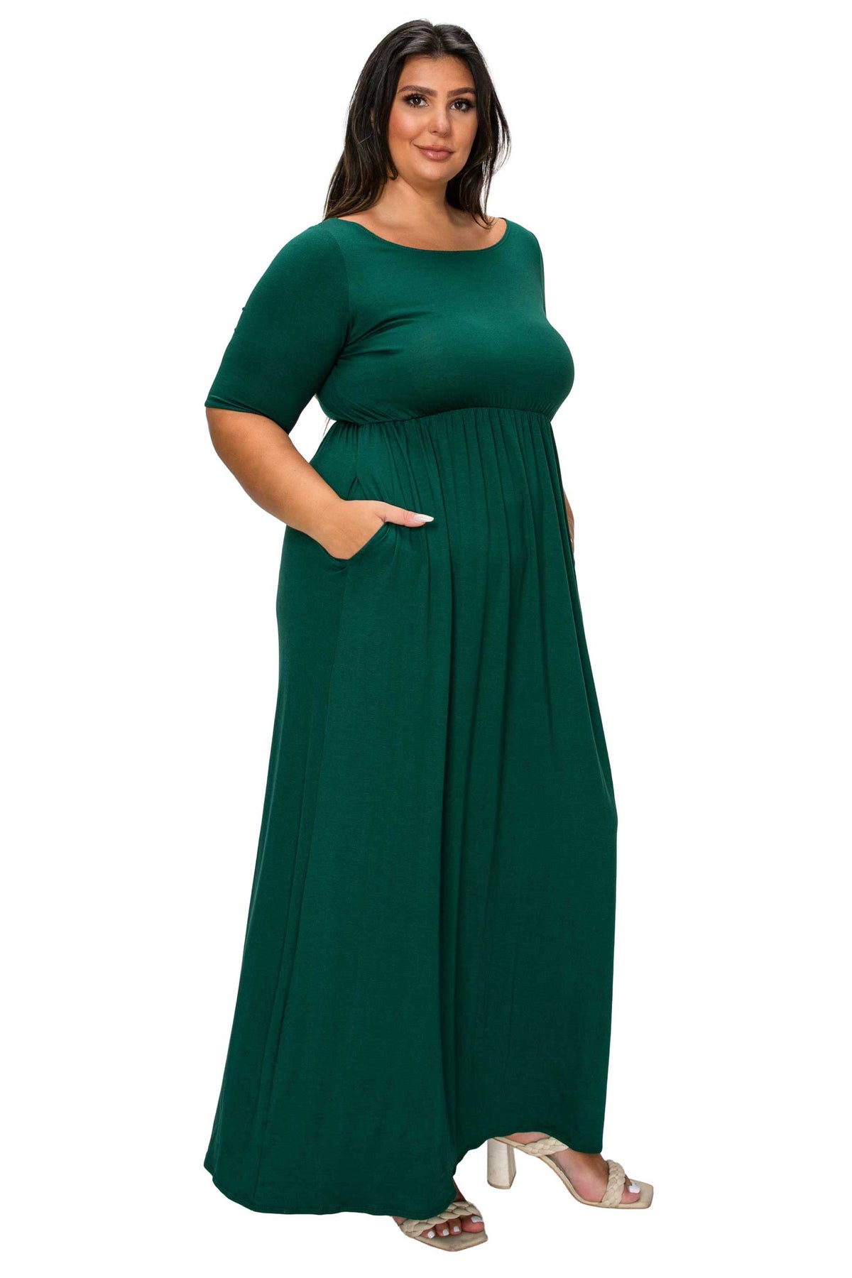 A woman modeling a dark green, short-sleeved maxi dress with a boat neckline and side pockets, paired with beige heeled sandals, against a plain white background.