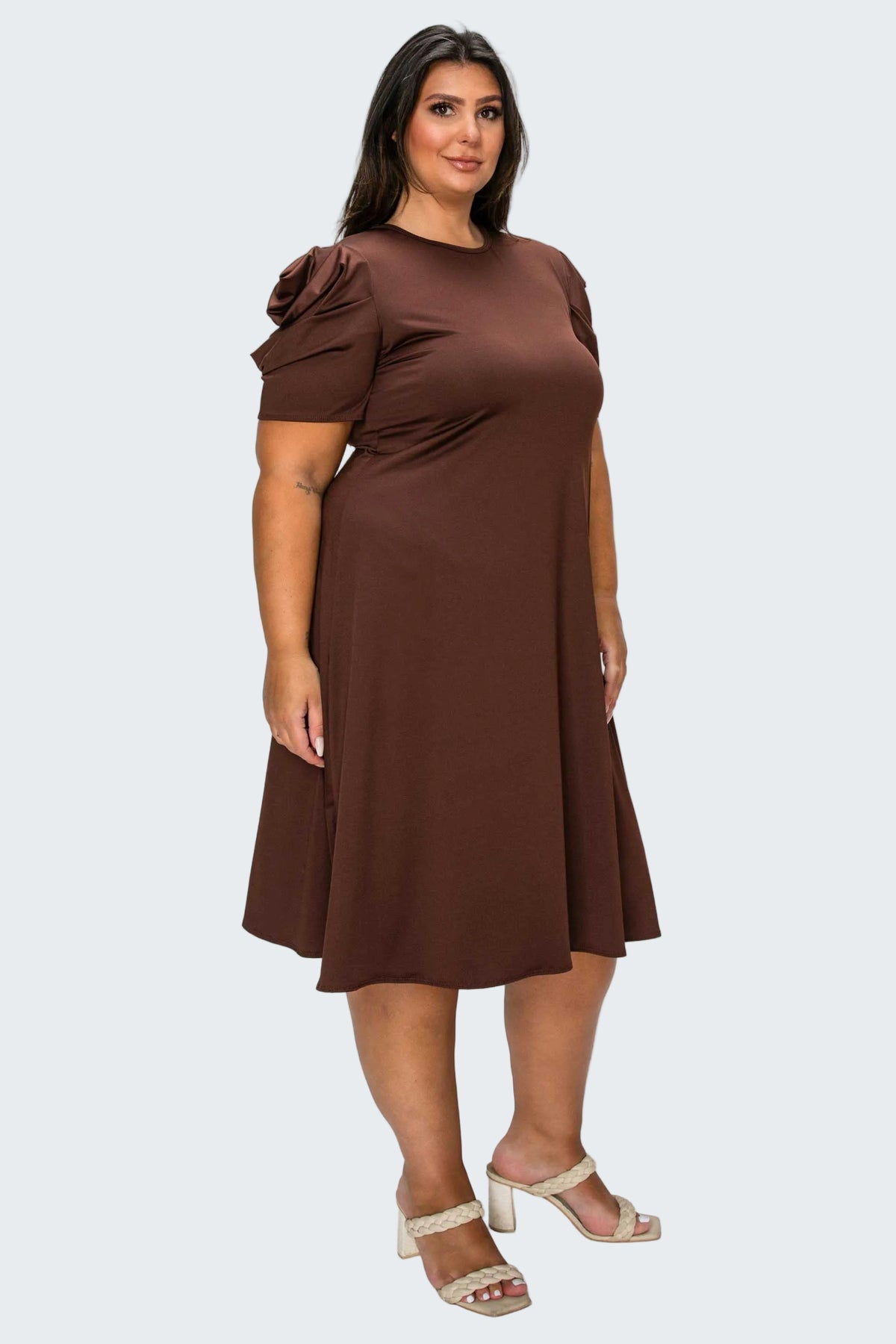 A woman modeling a brown short-sleeve dress with a ruffled shoulder detail, standing against a plain white background.