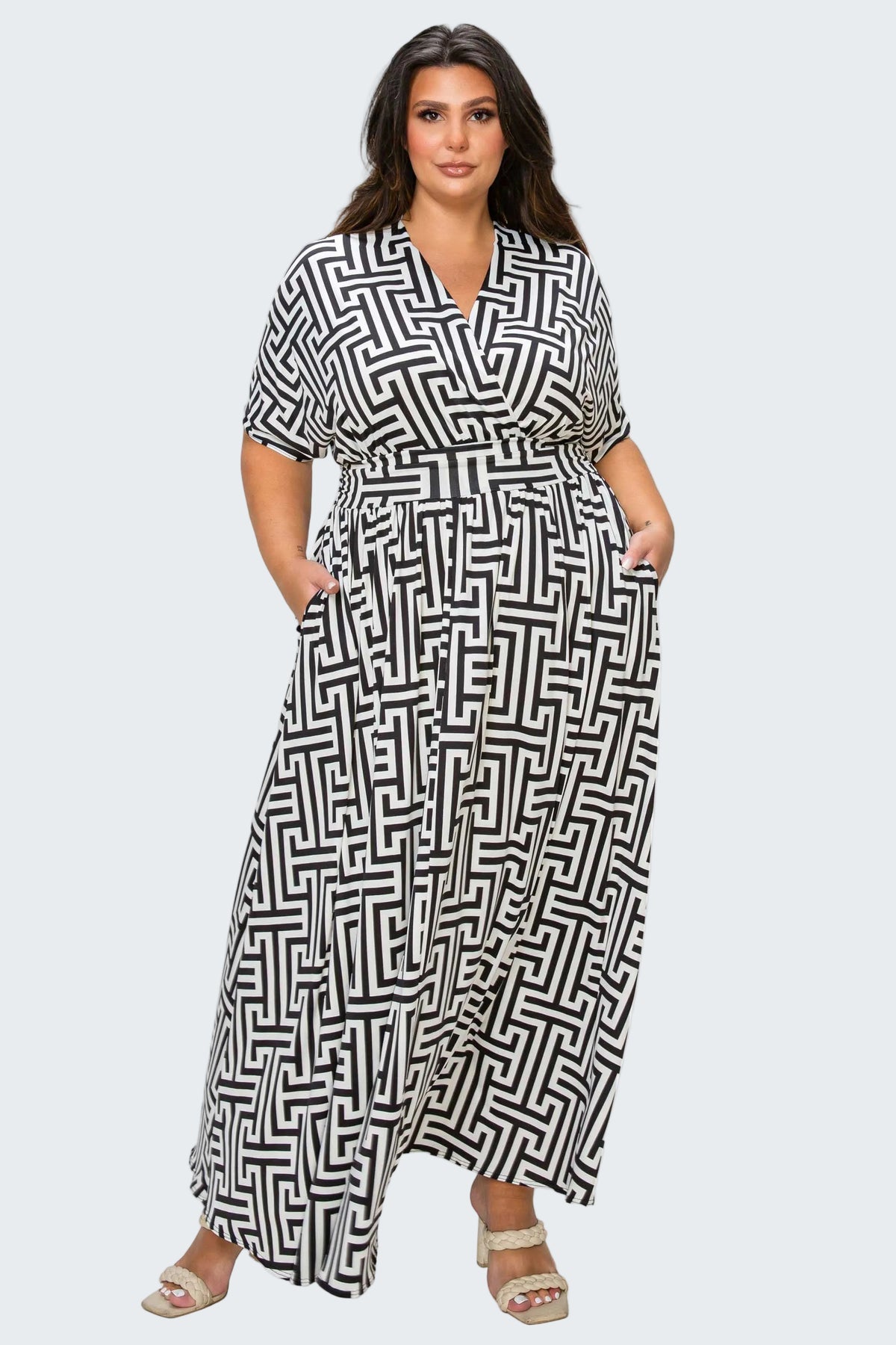 A woman modeling a black and white geometric maze-patterned maxi dress with short sleeves, a V-neckline, and a matching tie belt at the waist, set against a plain white background.