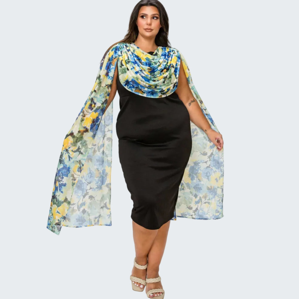 A woman modeling a black knee-length dress with a draped, semi-sheer blue, yellow, and green floral cape overlay, paired with beige platform sandals, against a plain white background.
