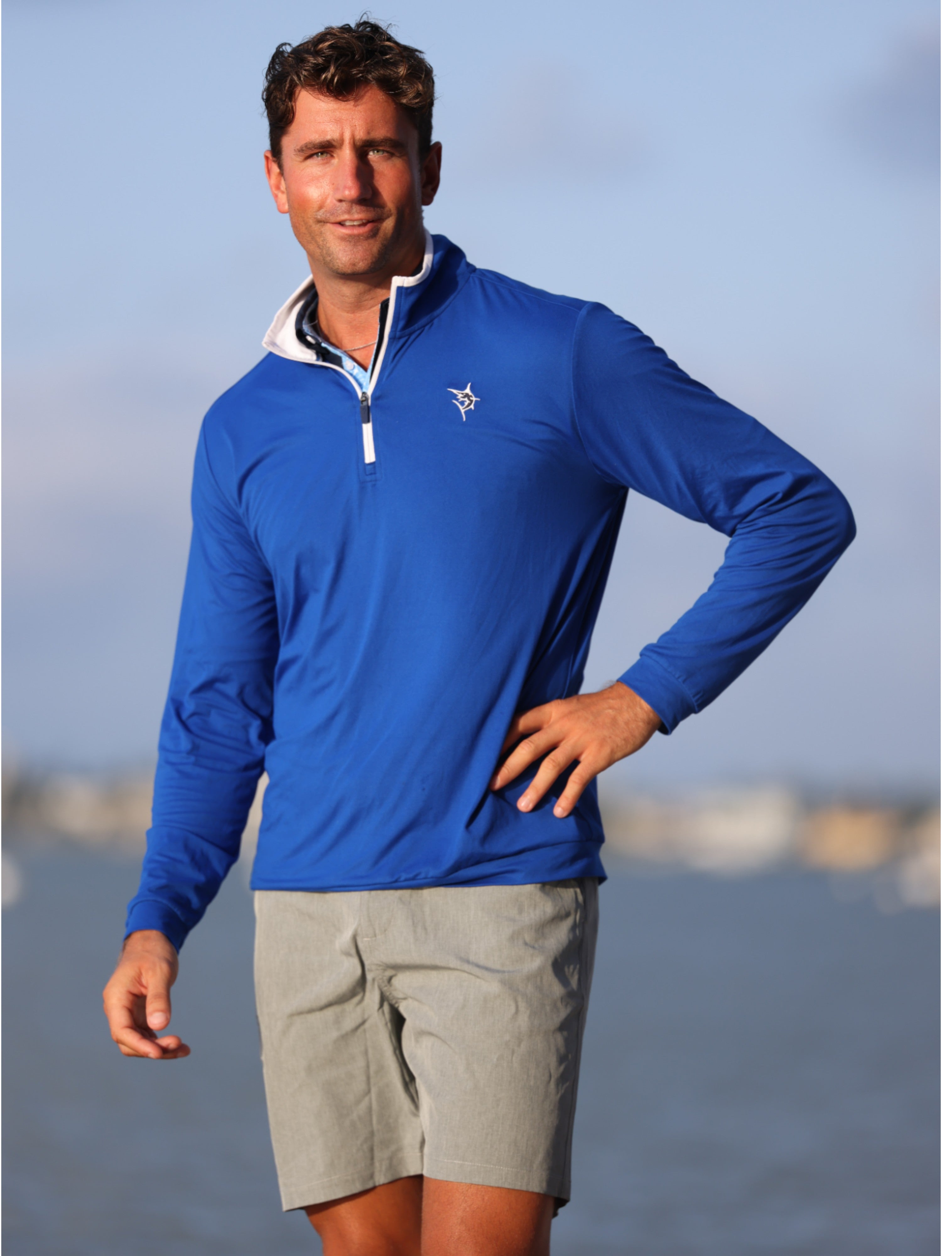 Man wearing a blue long-sleeve shirt with a logo, standing by water.