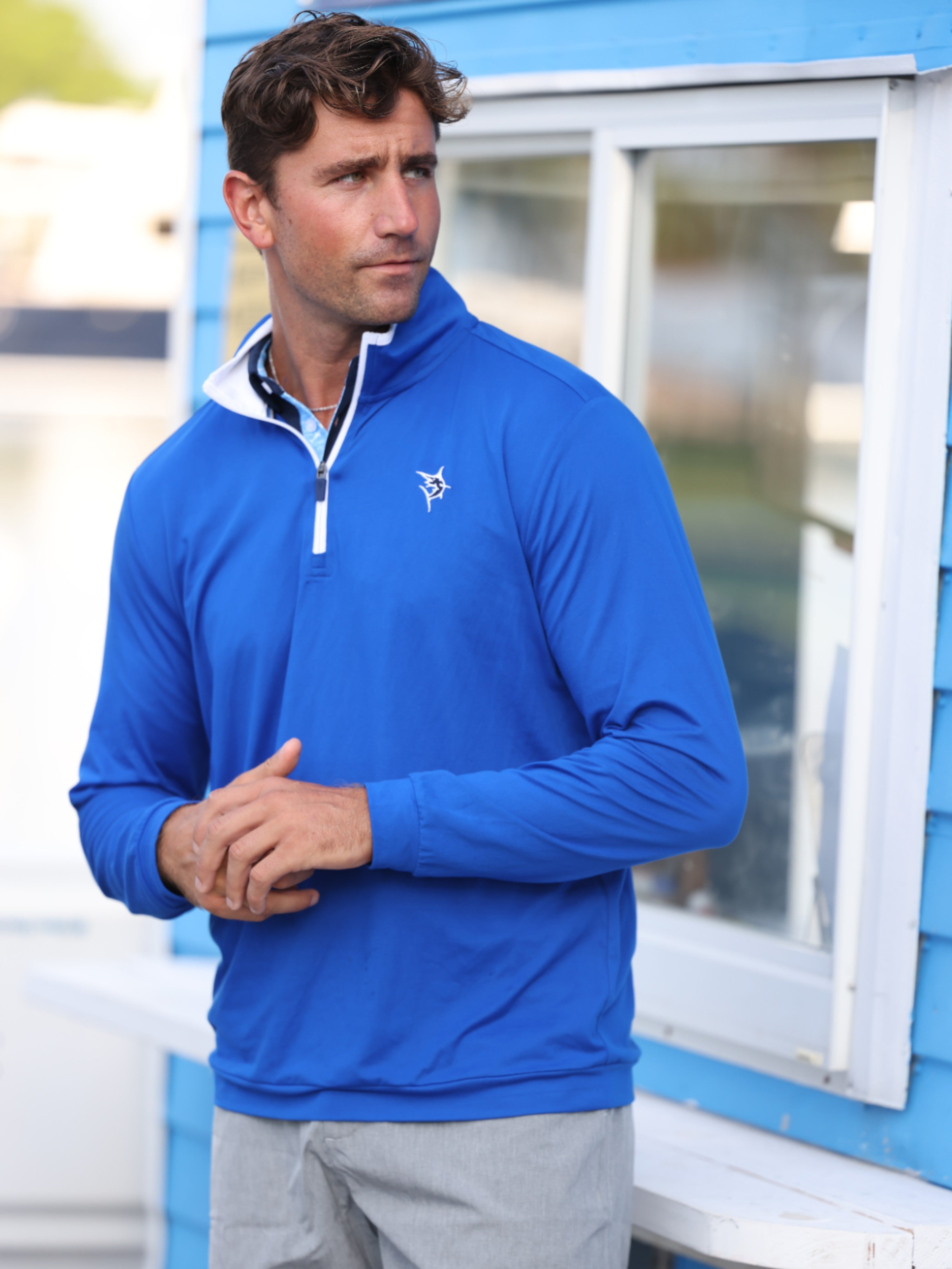 Man wearing a blue pullover with a logo, standing outdoors near a blue building.