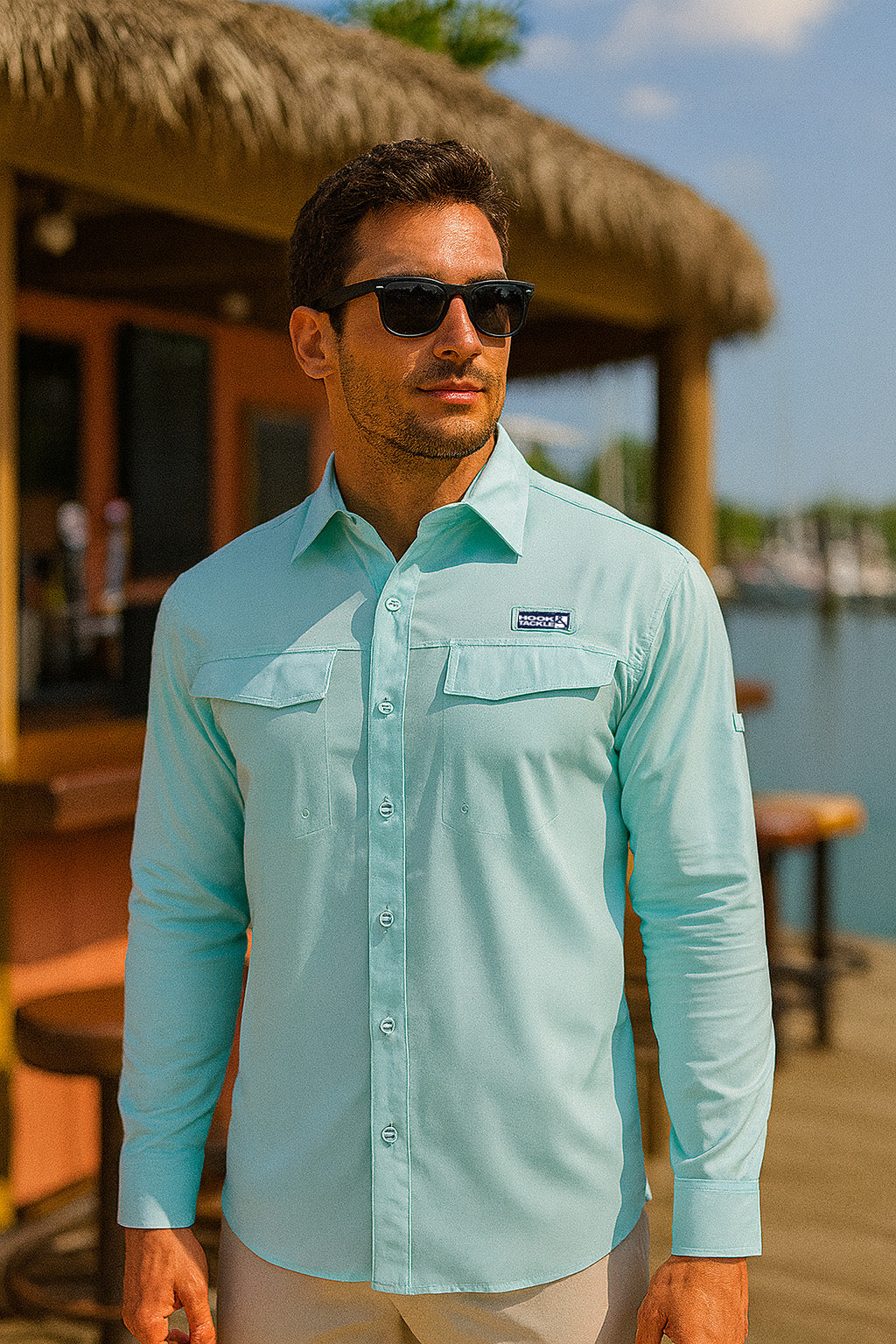 Man wearing a light blue shirt with a brand logo, standing on a wooden deck by a body of water.
