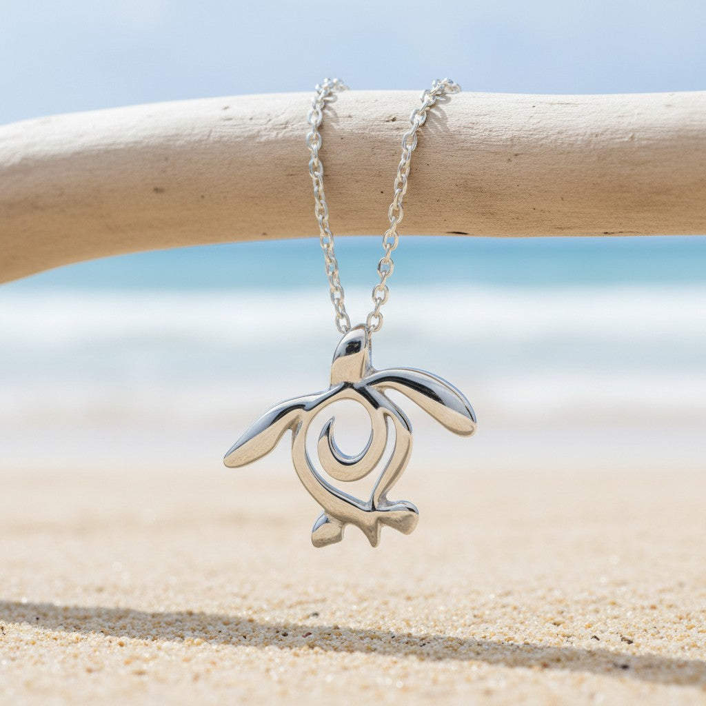 Silver turtle pendant necklace on a chain, displayed on a beach with sand and water in the background.