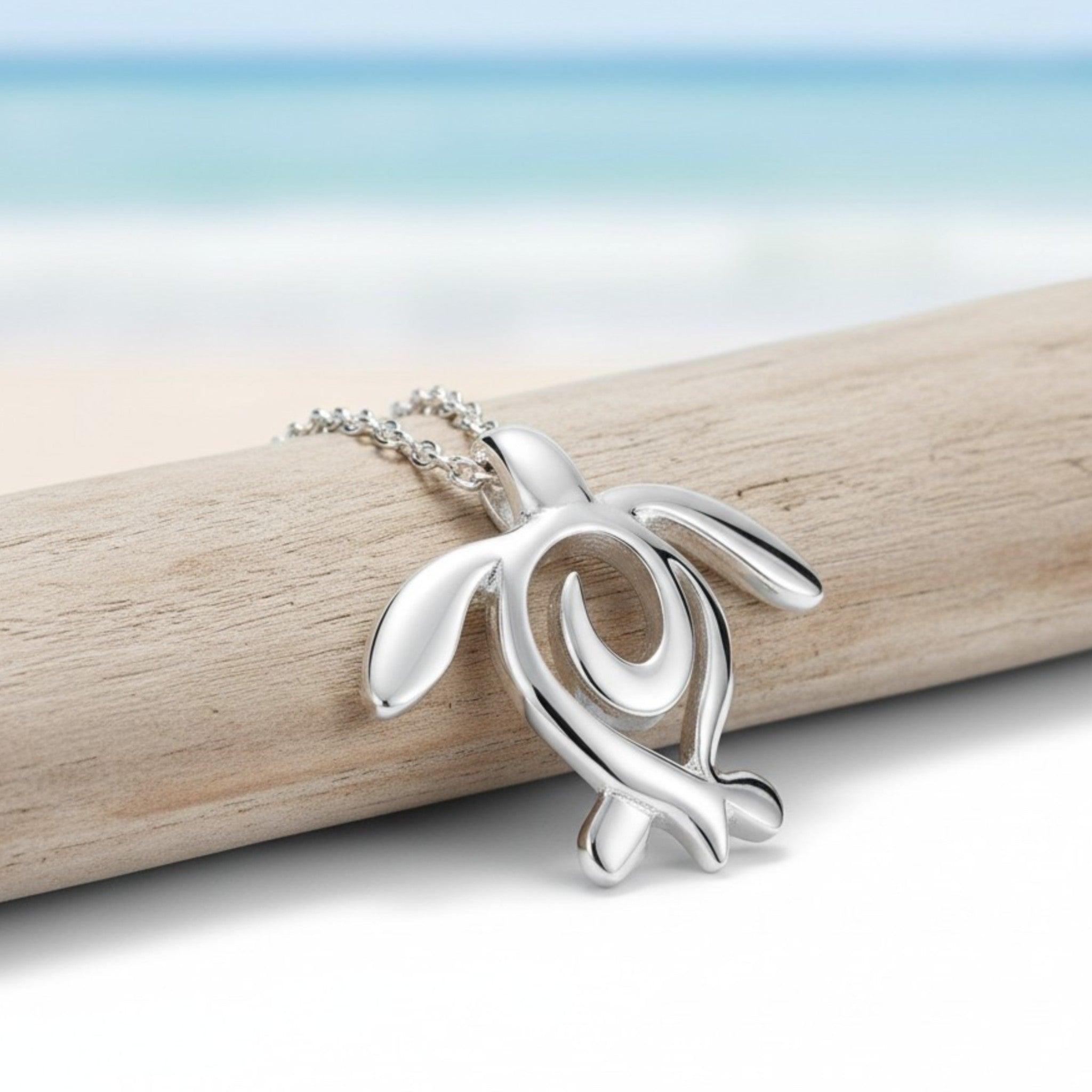 Silver turtle pendant on a chain resting on a piece of driftwood with a beach background.