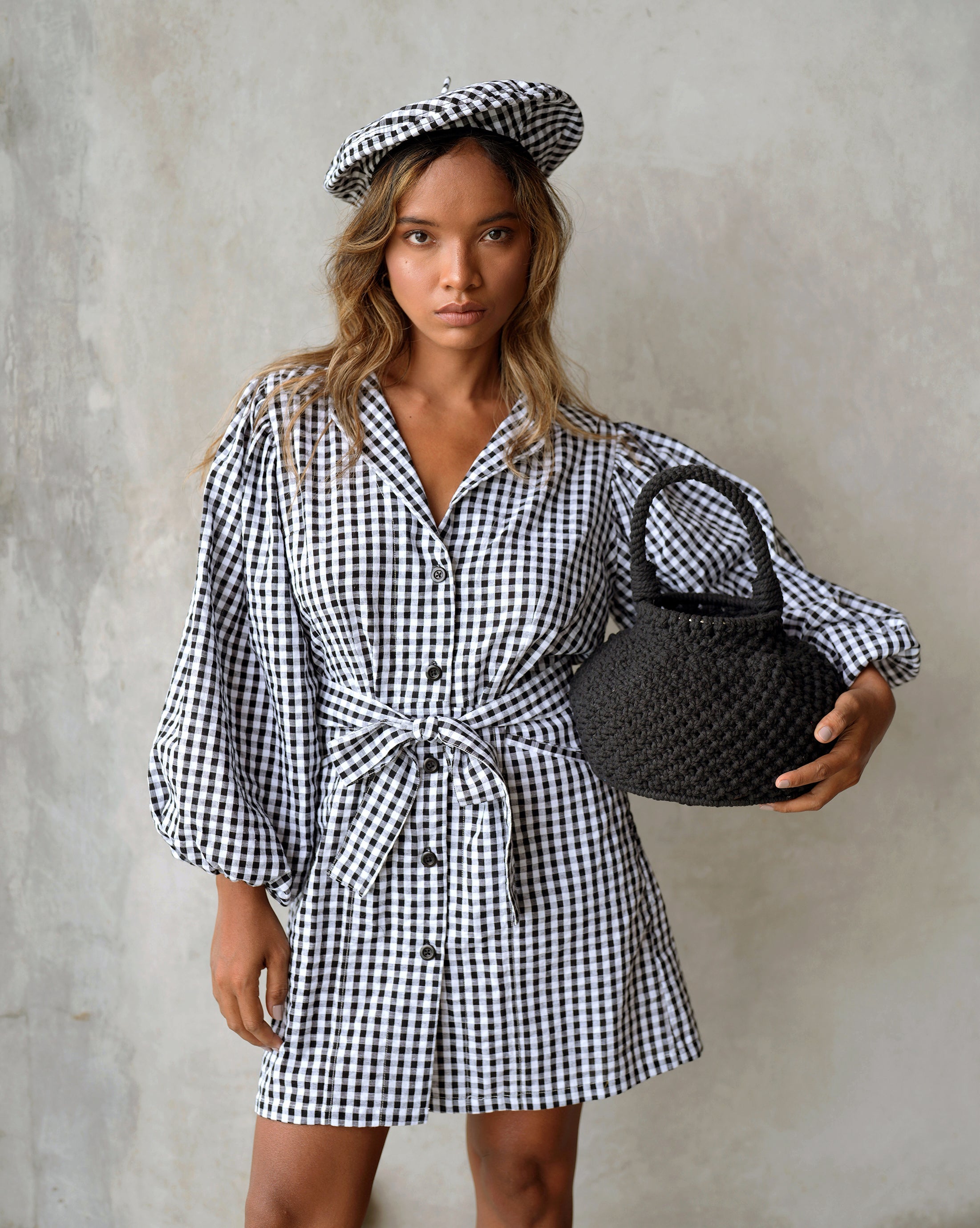 Woman wearing a black and white checkered dress holding a black handbag against a plain background