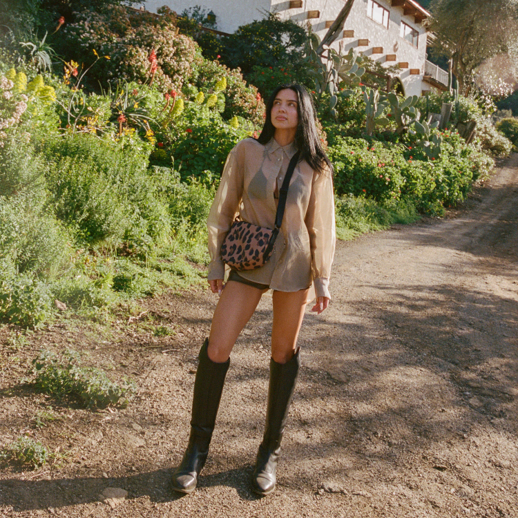Woman standing on a dirt path surrounded by greenery and a building in the background