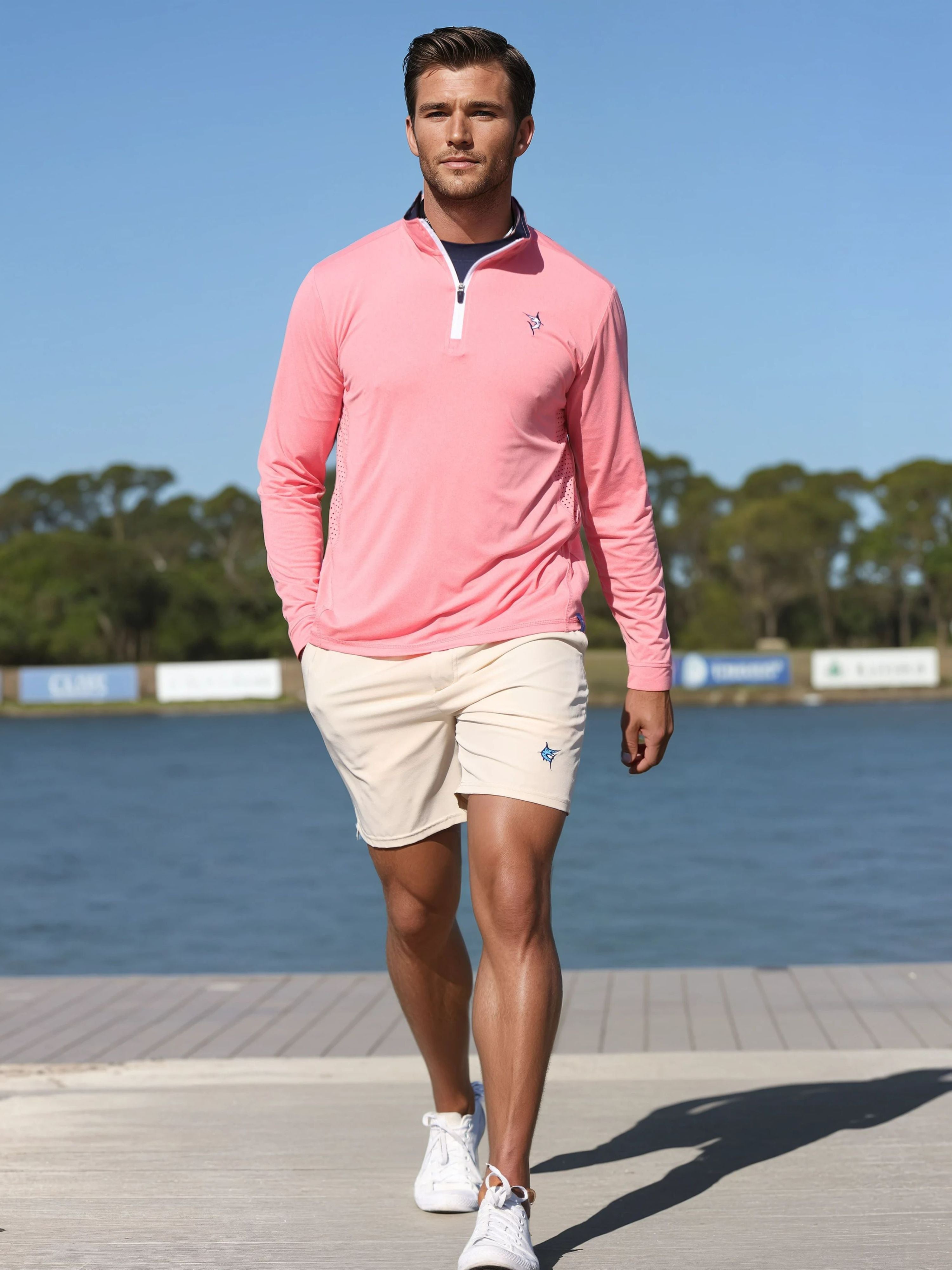 Man wearing a pink long-sleeve shirt and beige shorts standing on a dock by a body of water.