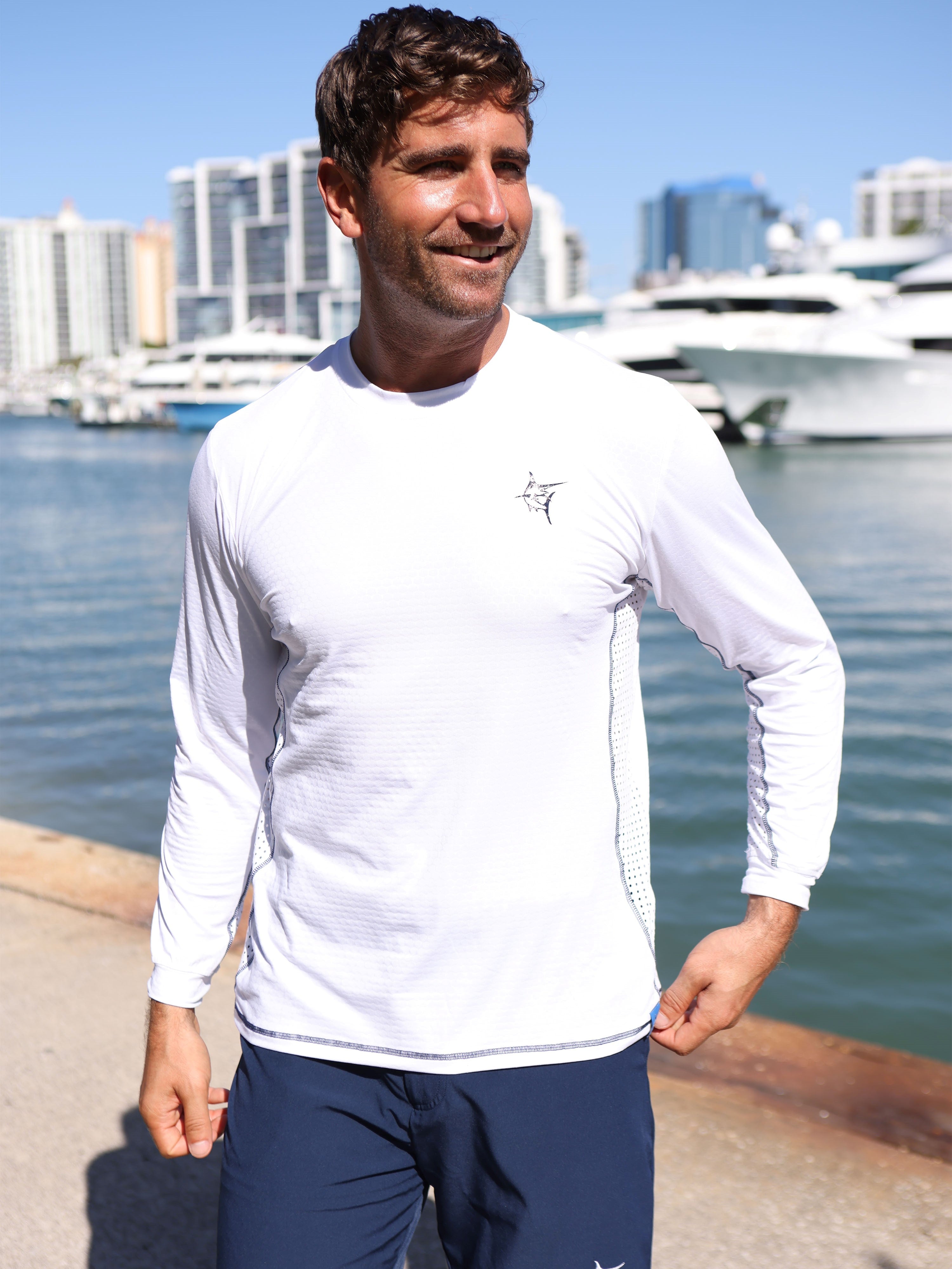 Man wearing a white long-sleeve shirt with a logo by a waterfront with boats and buildings in the background.