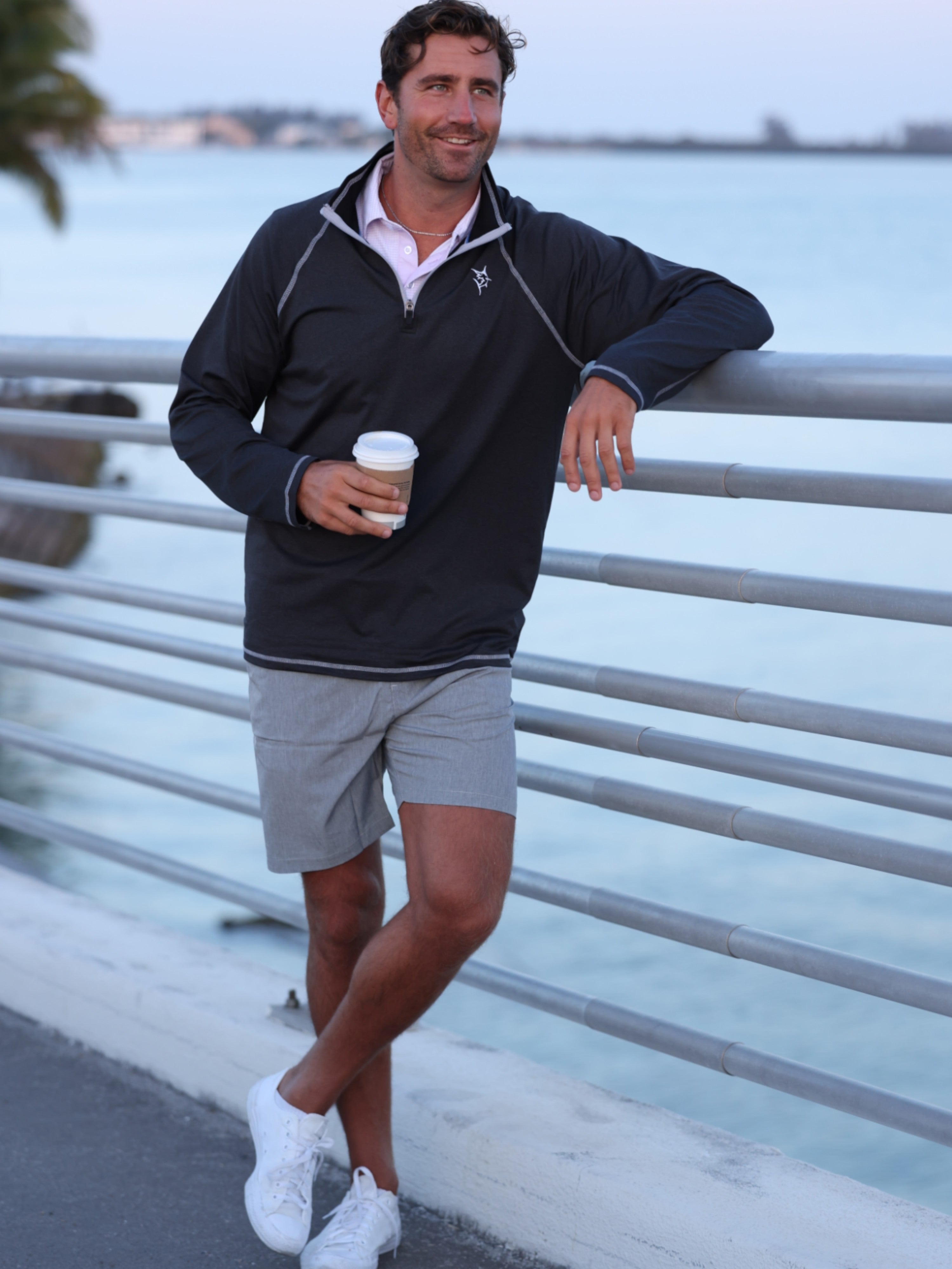 Man in navy jacket and gray shorts holding a coffee cup by a waterfront.