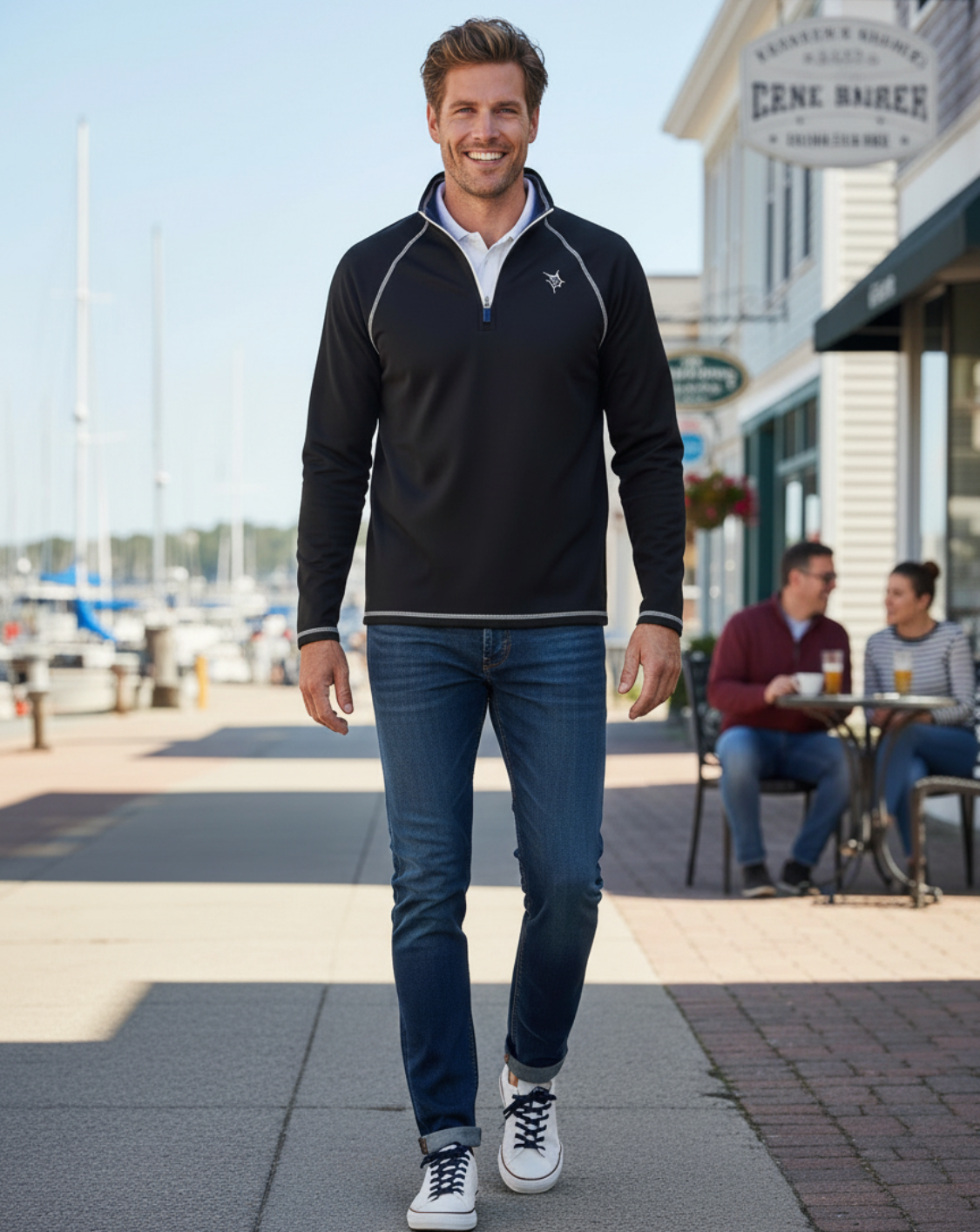 Man wearing a black quarter-zip sweatshirt and blue jeans standing outdoors near a marina.
