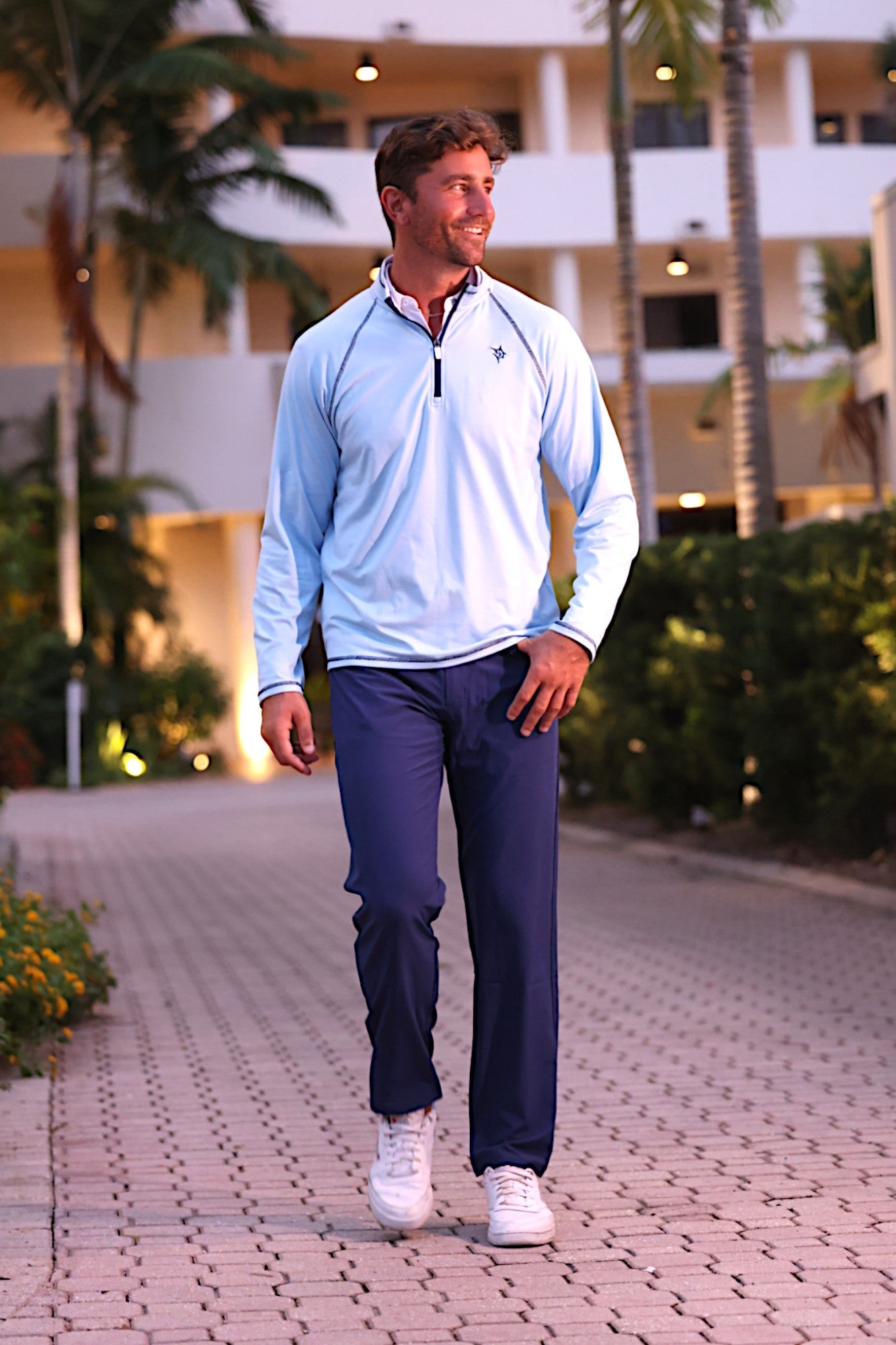 Man walking outdoors on a paved path with palm trees and a building in the background