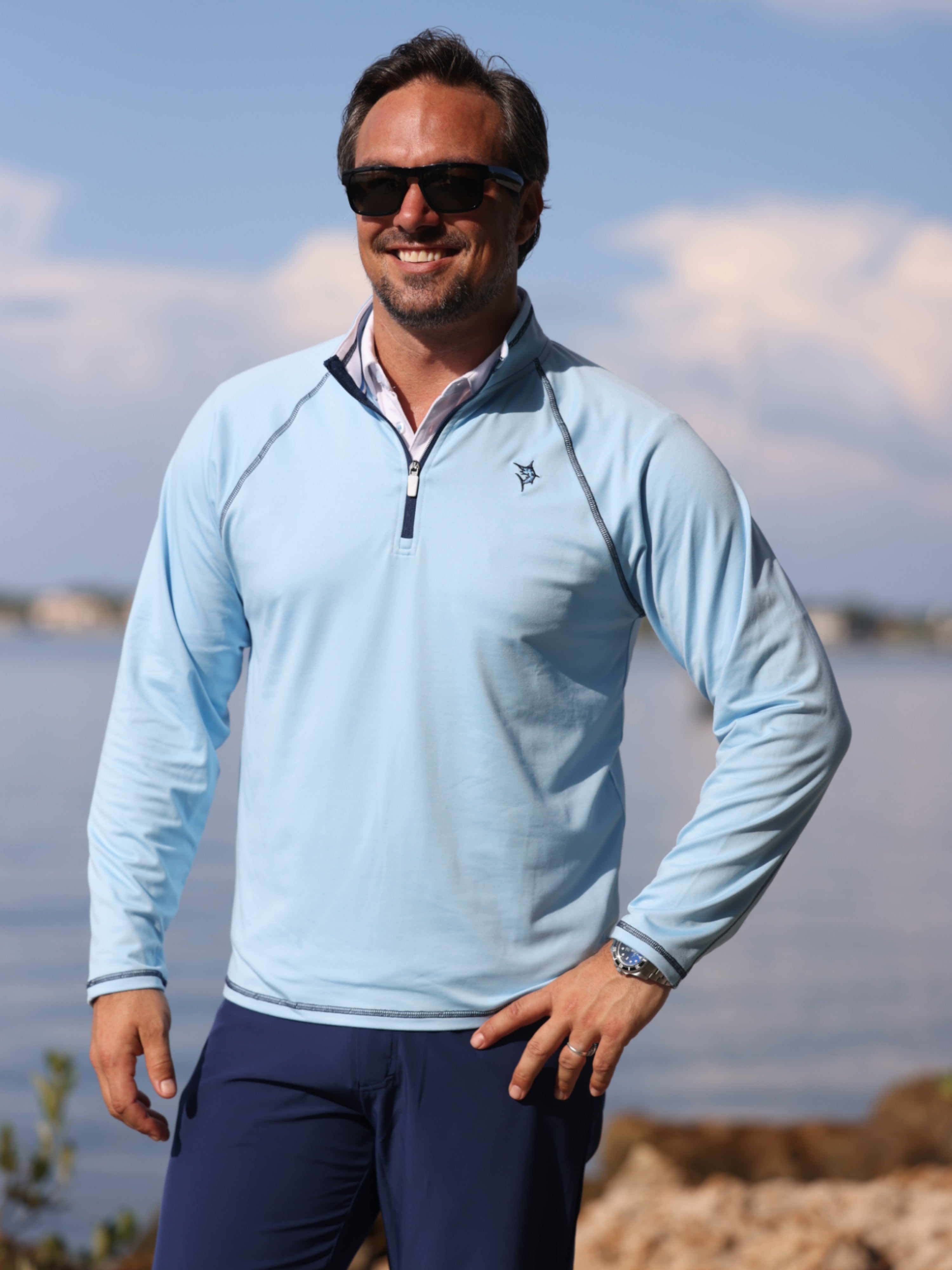 Man wearing a light blue long-sleeve shirt and dark sunglasses standing by a body of water.