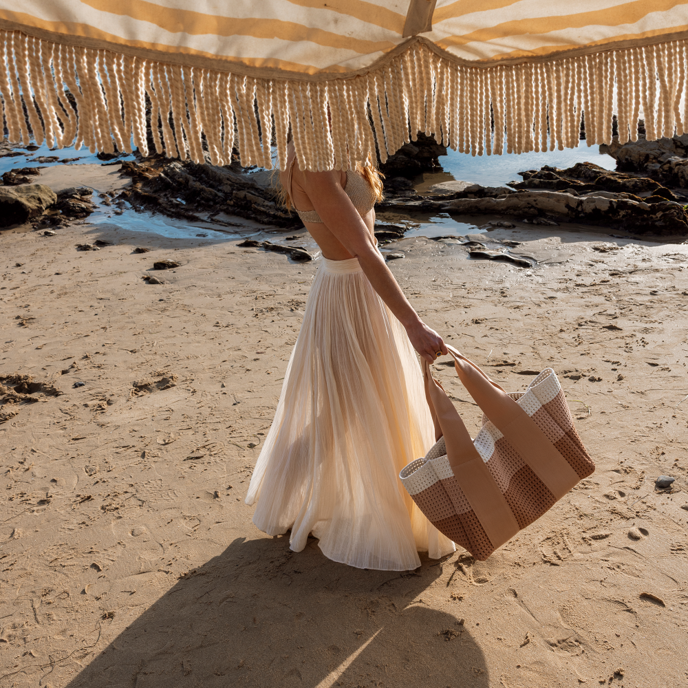 Woman in a white dress holding a woven bag on a sandy beach with a striped canopy.