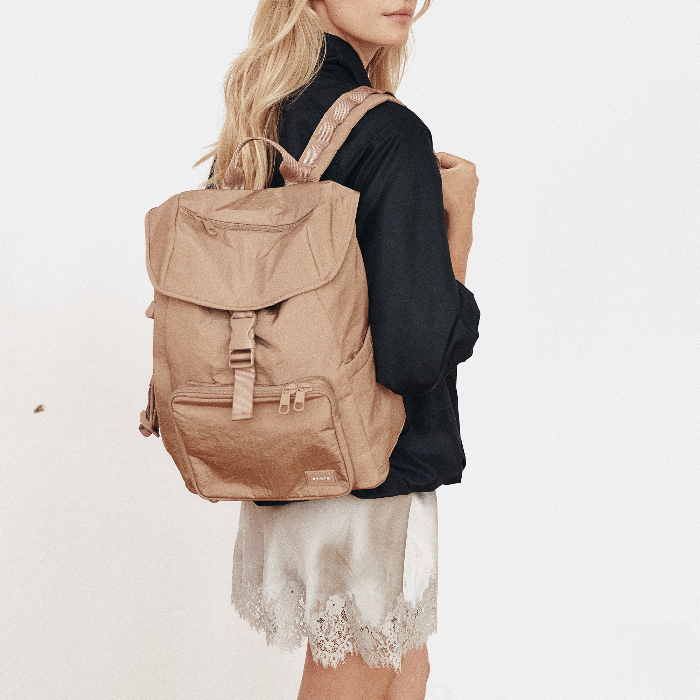 A woman wearing a black long-sleeve top and a light beige satin skirt with lace trim, carrying a beige canvas backpack on her shoulder against a plain white background.