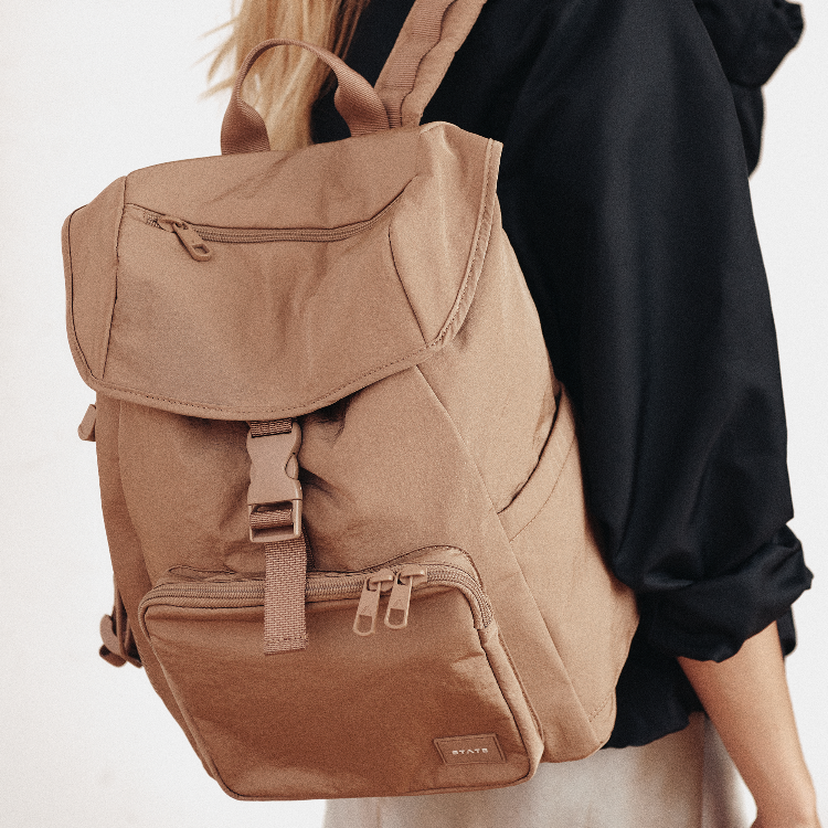 A tan canvas backpack with multiple zippered pockets and a buckle strap, shown worn by a person in a black top against a clean white background.