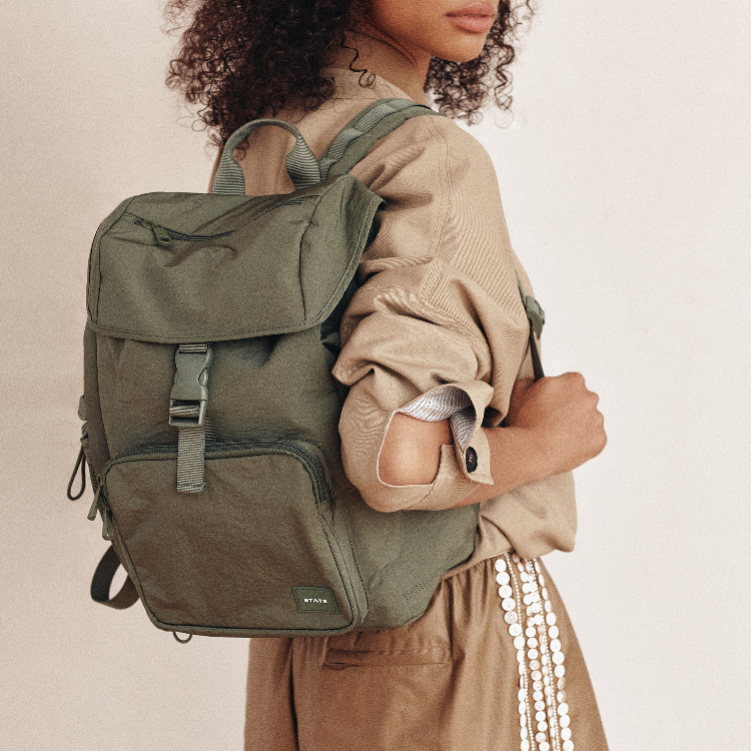 An olive green canvas backpack with multiple zippered compartments and a buckle strap, shown on a model wearing a beige shirt and brown skirt against a plain beige background.