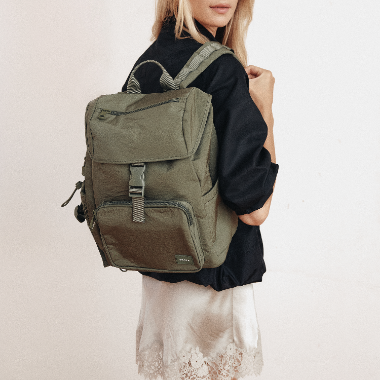 An olive green backpack with gray straps and buckles, worn by a model wearing a black shirt and a white lace skirt against a plain off-white background.