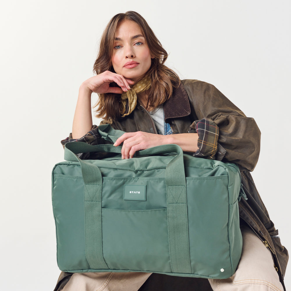 Woman holding a green duffel bag with a visible brand logo on a plain background
