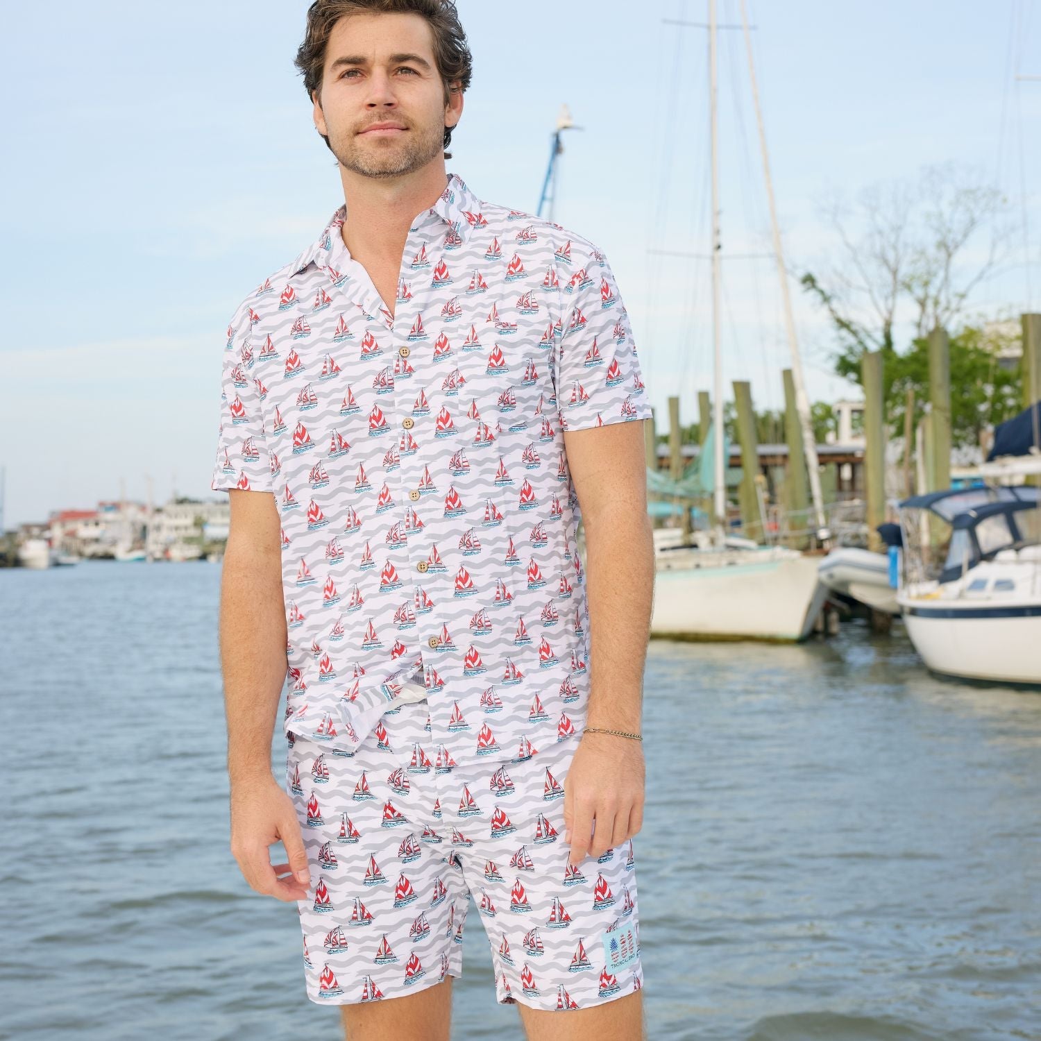 Man wearing a patterned shirt and shorts by a marina with boats and water in the background.