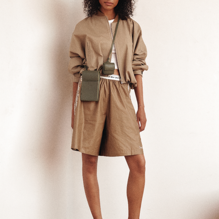 A model wearing a beige cropped bomber jacket and matching beige shorts, styled with olive green crossbody and belt bags, standing against a plain off-white wall.