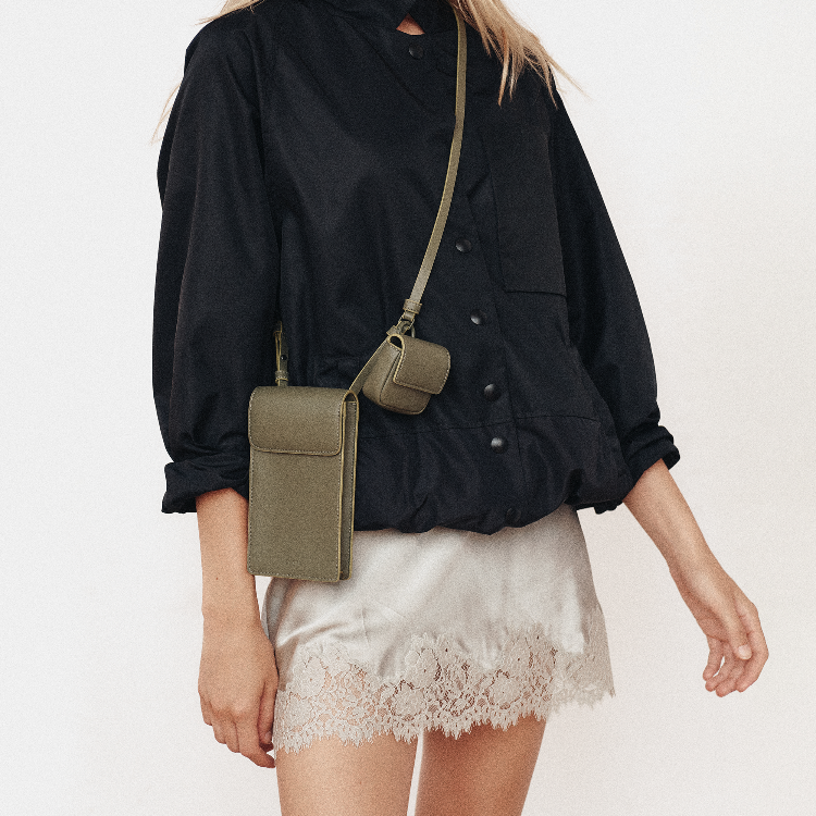 A model wearing a black button-up shirt and a white lace-trimmed skirt, carrying an olive green crossbody phone pouch bag with a matching small pouch on the back.