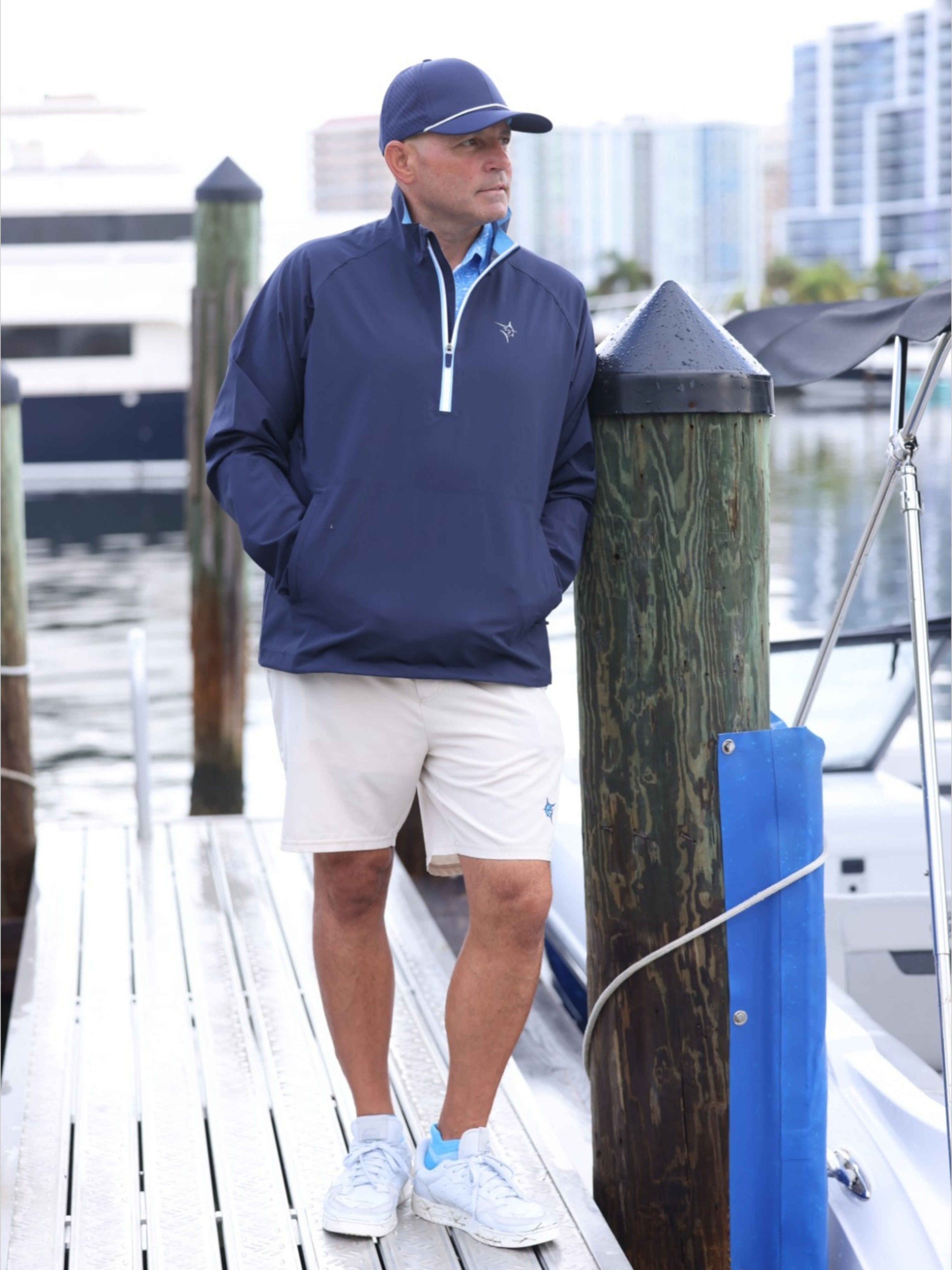 Man in navy jacket and white shorts standing on a dock with boats and buildings in the background