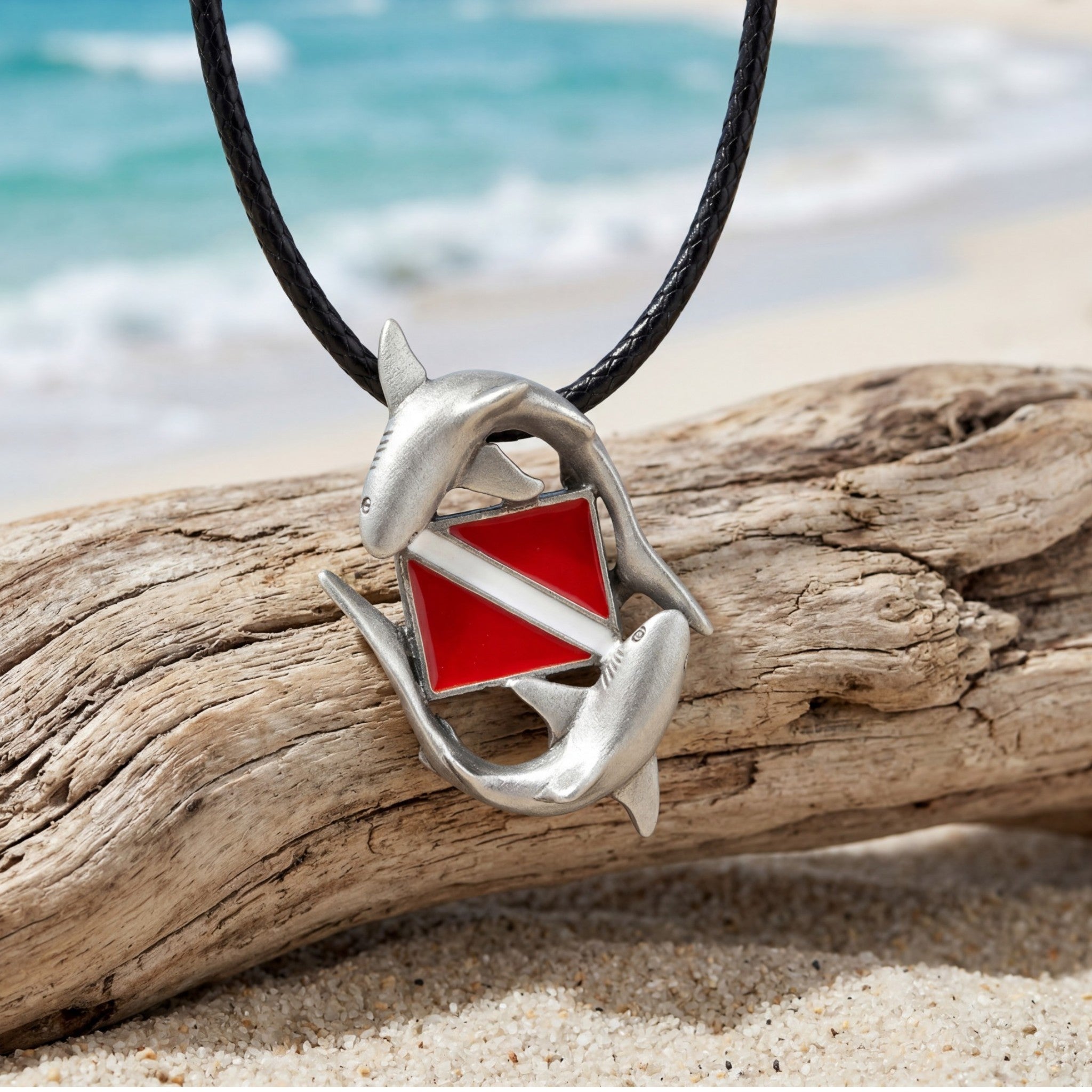 Shark pendant with red square on a black cord, placed on driftwood with ocean background