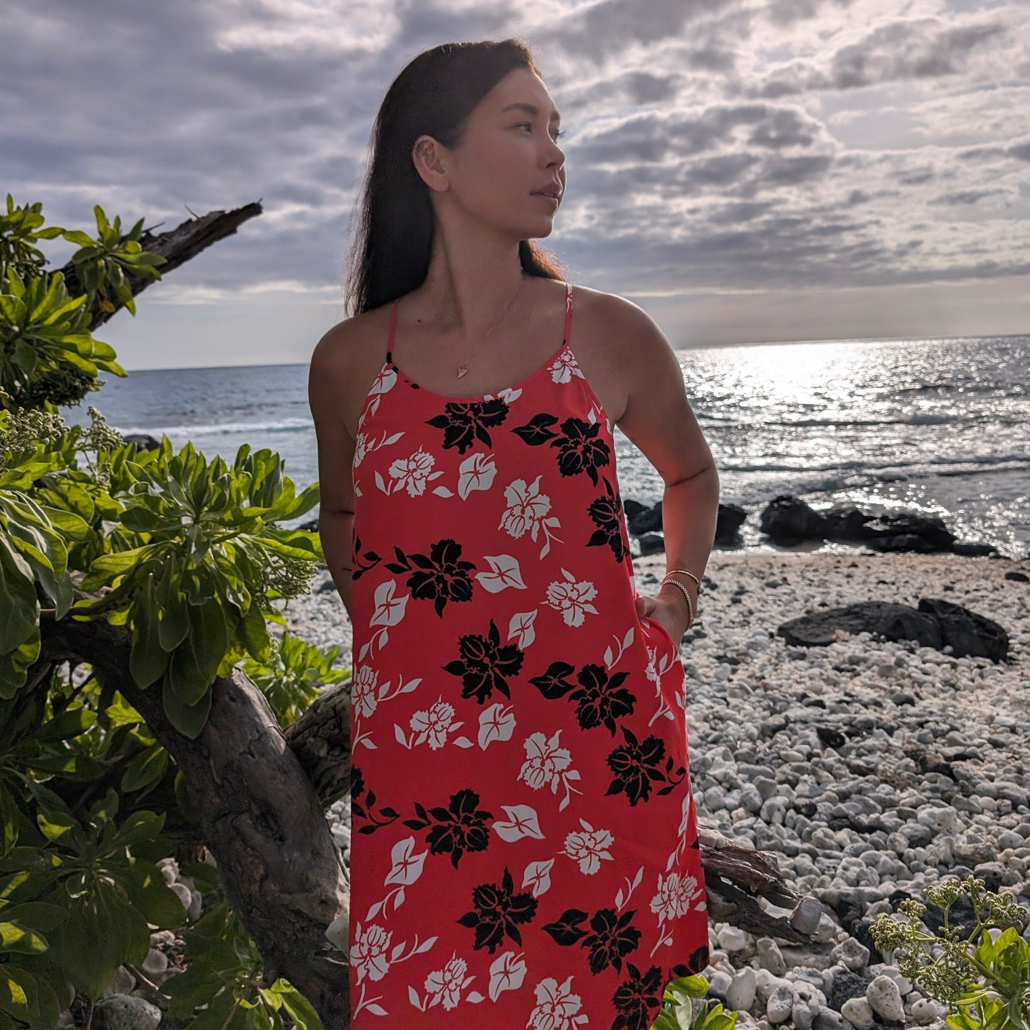 Woman in a red floral dress standing on a rocky beach with ocean and sky in the background