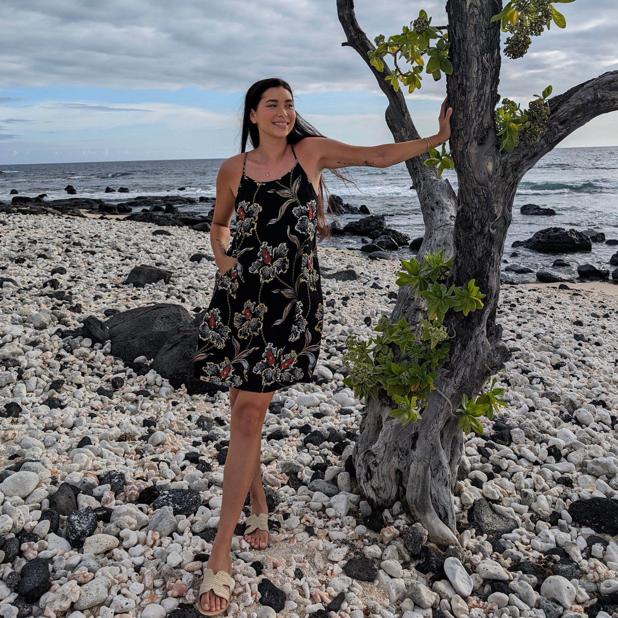 Woman in a black floral dress standing on a rocky beach with ocean and sky in the background