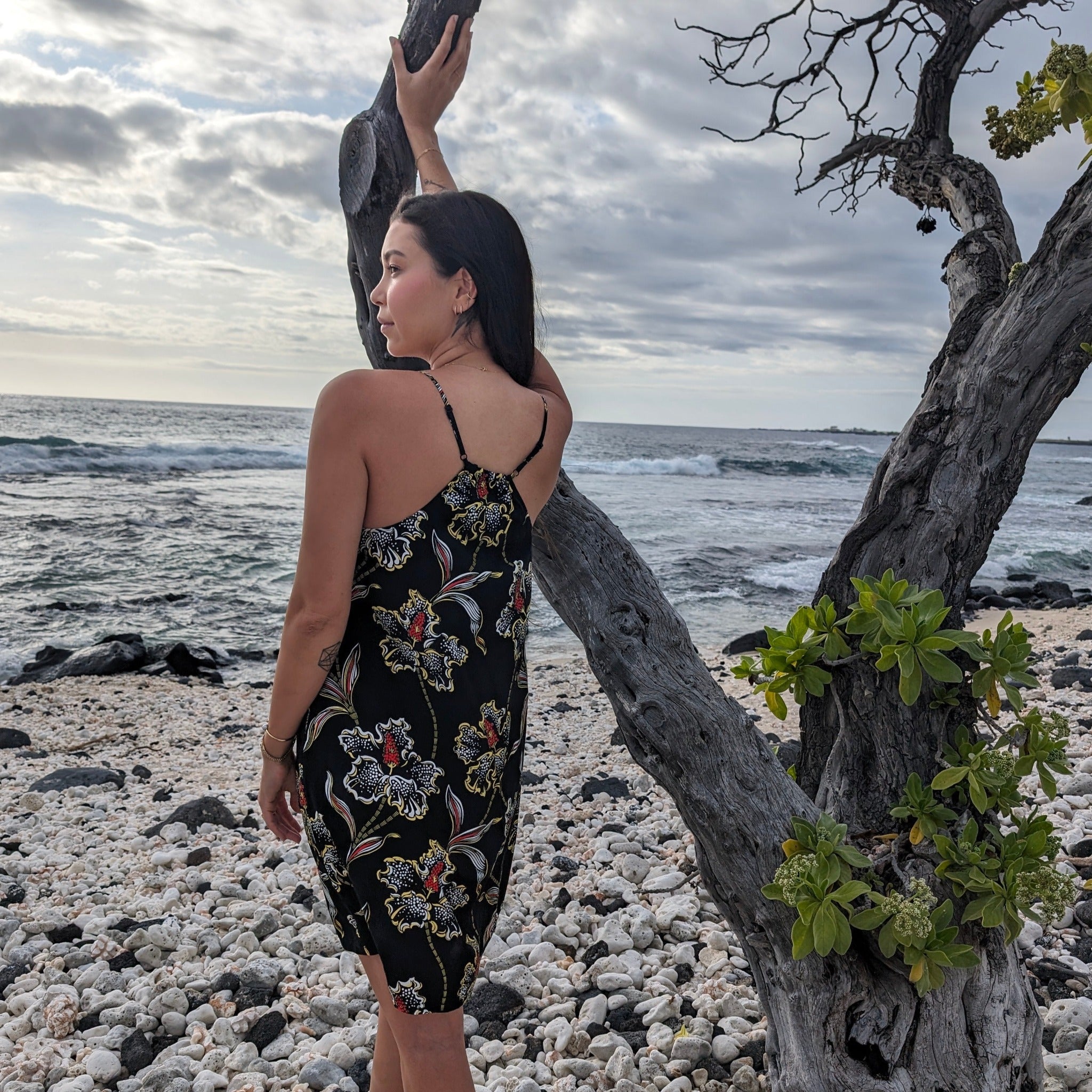 Woman in a floral dress standing on a rocky beach with ocean and cloudy sky in the background