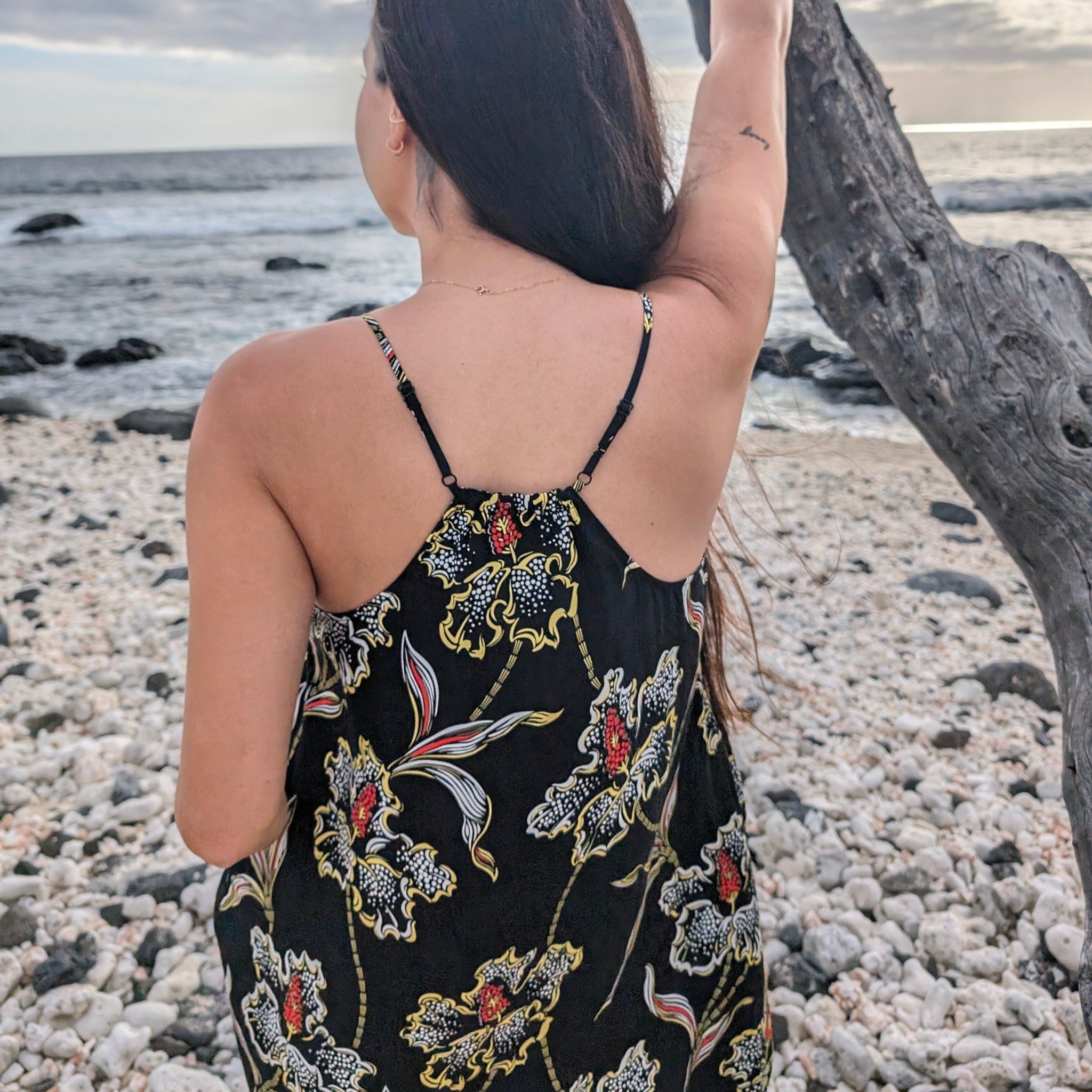 Woman in a black floral dress standing on a pebbly beach with ocean in the background