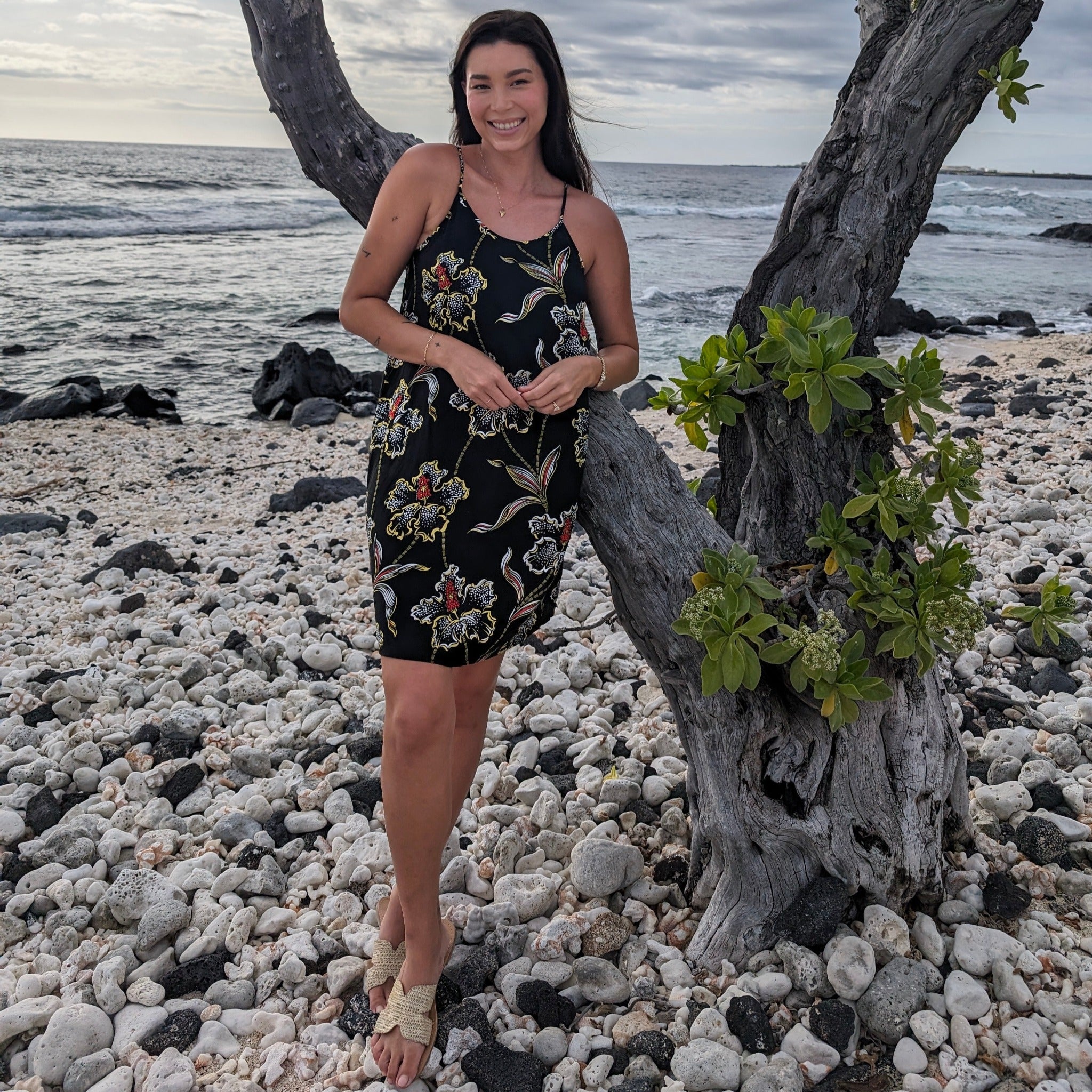 Woman in a floral dress standing on a pebbly beach with ocean and sky in the background