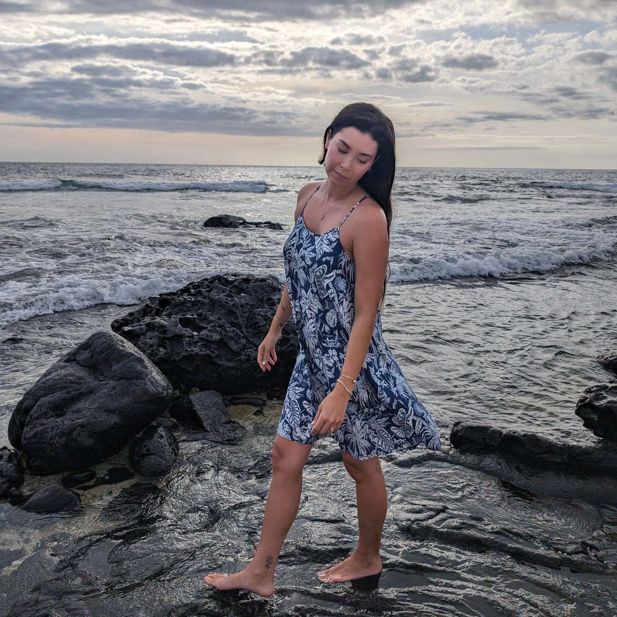 Woman in a blue floral dress standing on a rocky beach with ocean waves in the background.