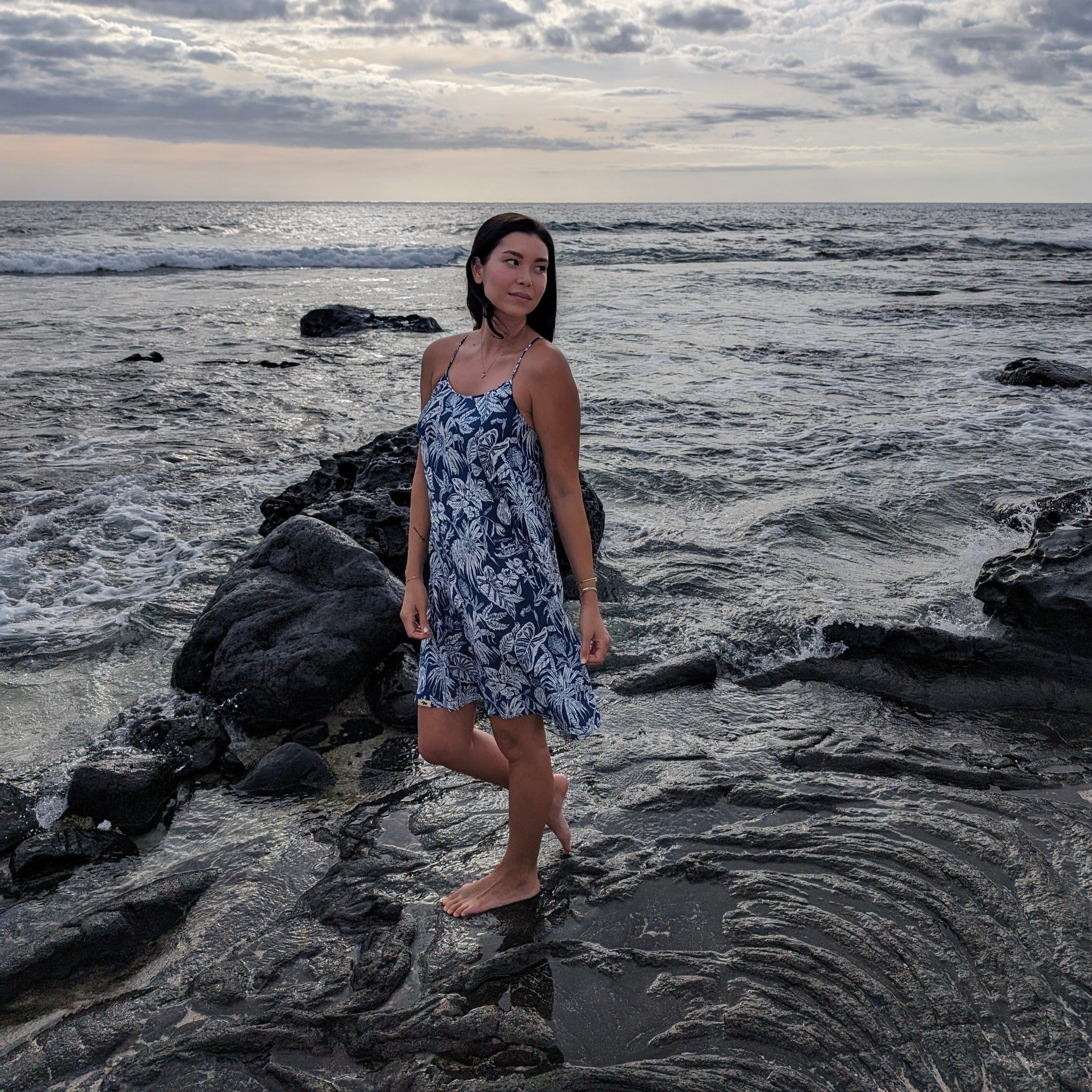 Woman in a blue floral dress standing on a rocky beach with ocean waves in the background.