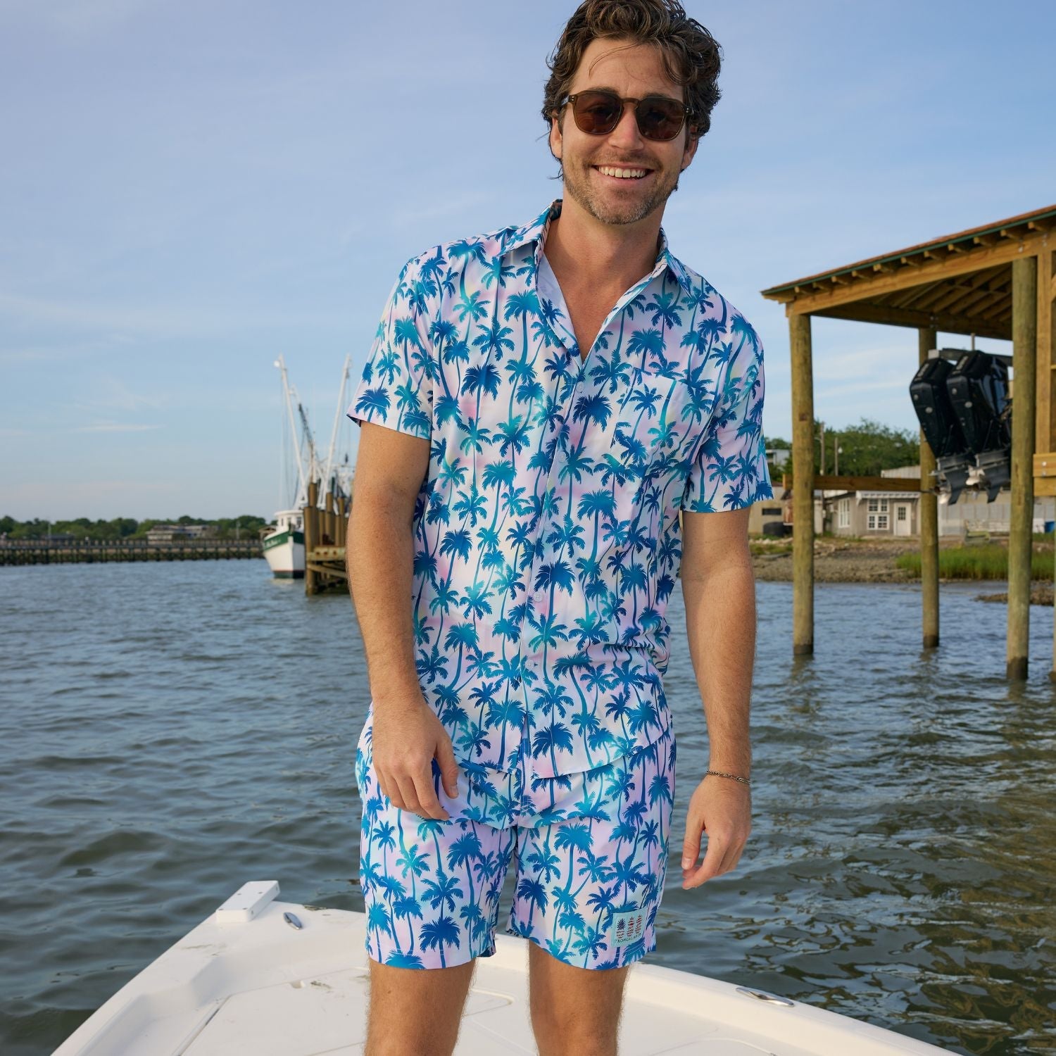 Man wearing a blue palm tree patterned shirt and shorts on a boat by water.
