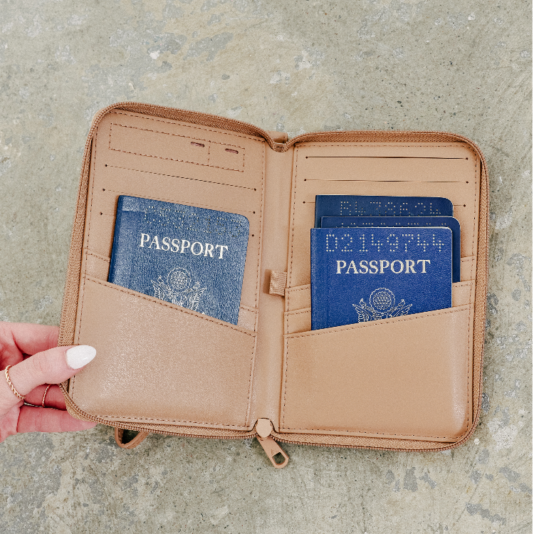 A tan faux-leather passport holder wallet shown open, held by a hand with white nail polish on a textured gray stone surface. The holder contains blue passports and cards.