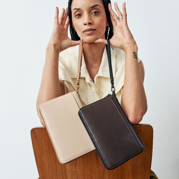 A woman holds two leather folio-style crossbody bags against a light gray background, one in beige and one in dark brown. She is wearing a cream blouse and has her hands raised near her face.